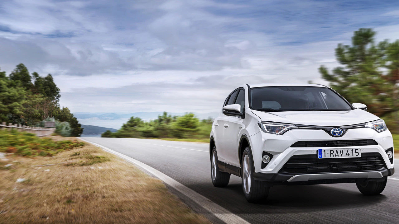 Ein weißer SUV fährt auf einer Landstraße durch eine grüne, bewaldete Landschaft mit bewölktem Himmel im Hintergrund.
