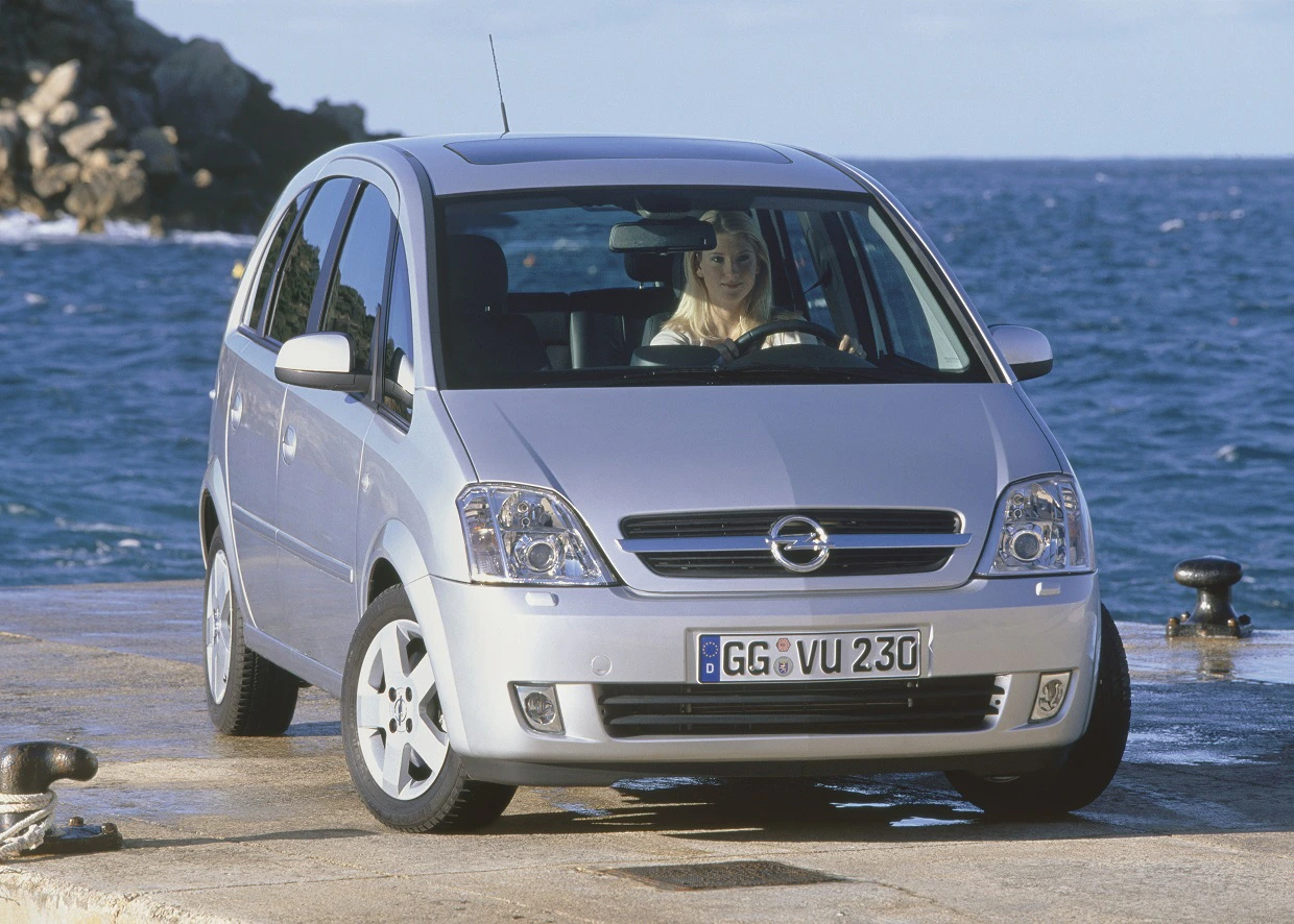 Silberner Opel Meriva parkt auf einem Pier mit Blick auf das Meer. Im Auto sitzt eine Person am Steuer. Im Hintergrund sind Wellen sichtbar.