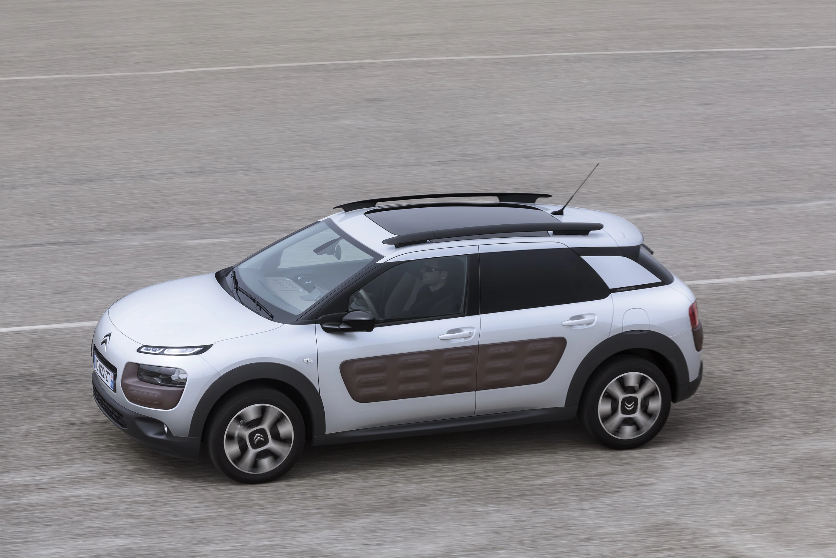 Graues Citroën-Auto mit braunen Akzenten und Dachträgern, fährt auf einer grauen Straße. Ansicht von oben.