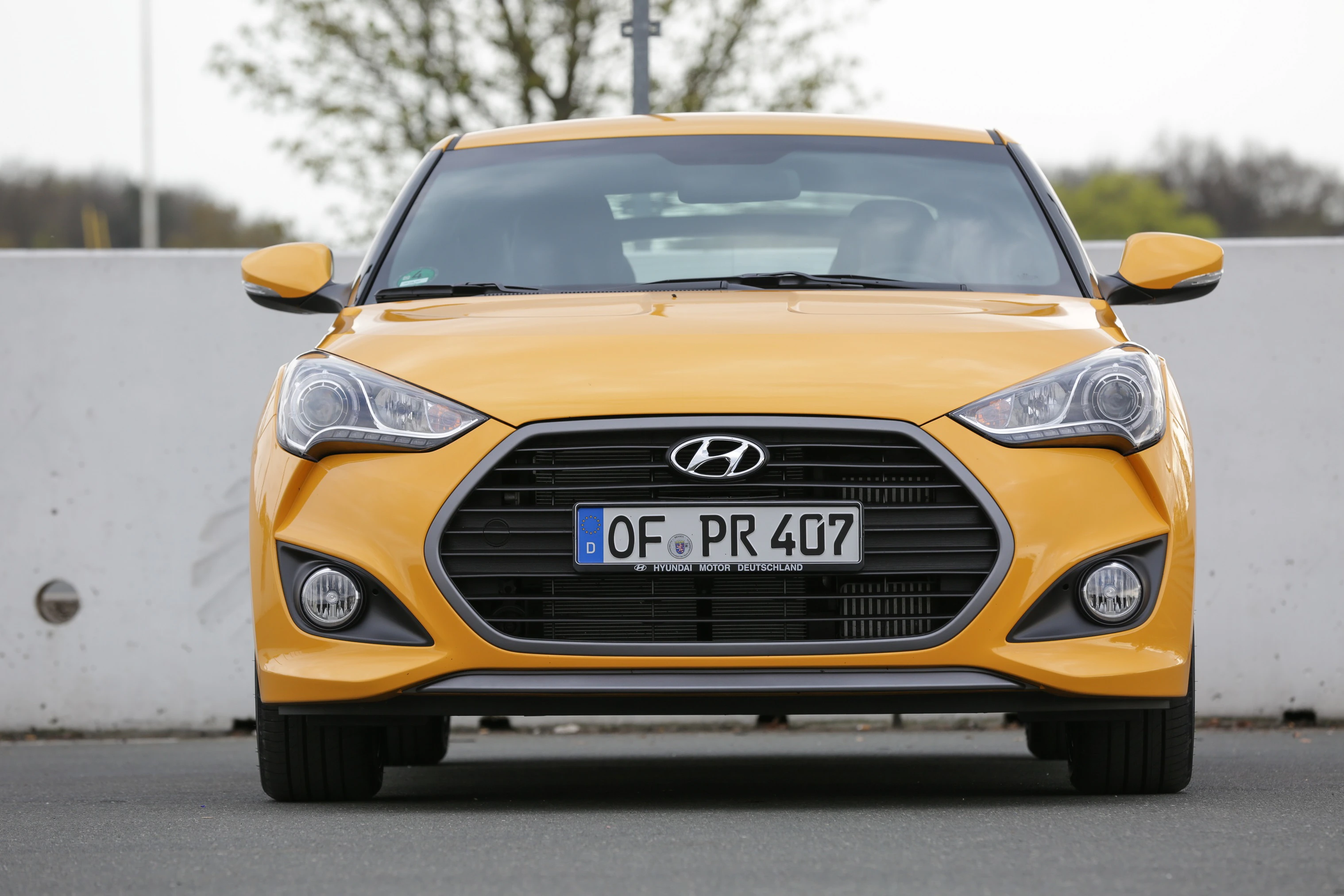 Gelber Hyundai Veloster von vorne, geparkt vor einer weißen Betonwand auf einem Parkplatz, umgeben von unscharfer Vegetation im Hintergrund.