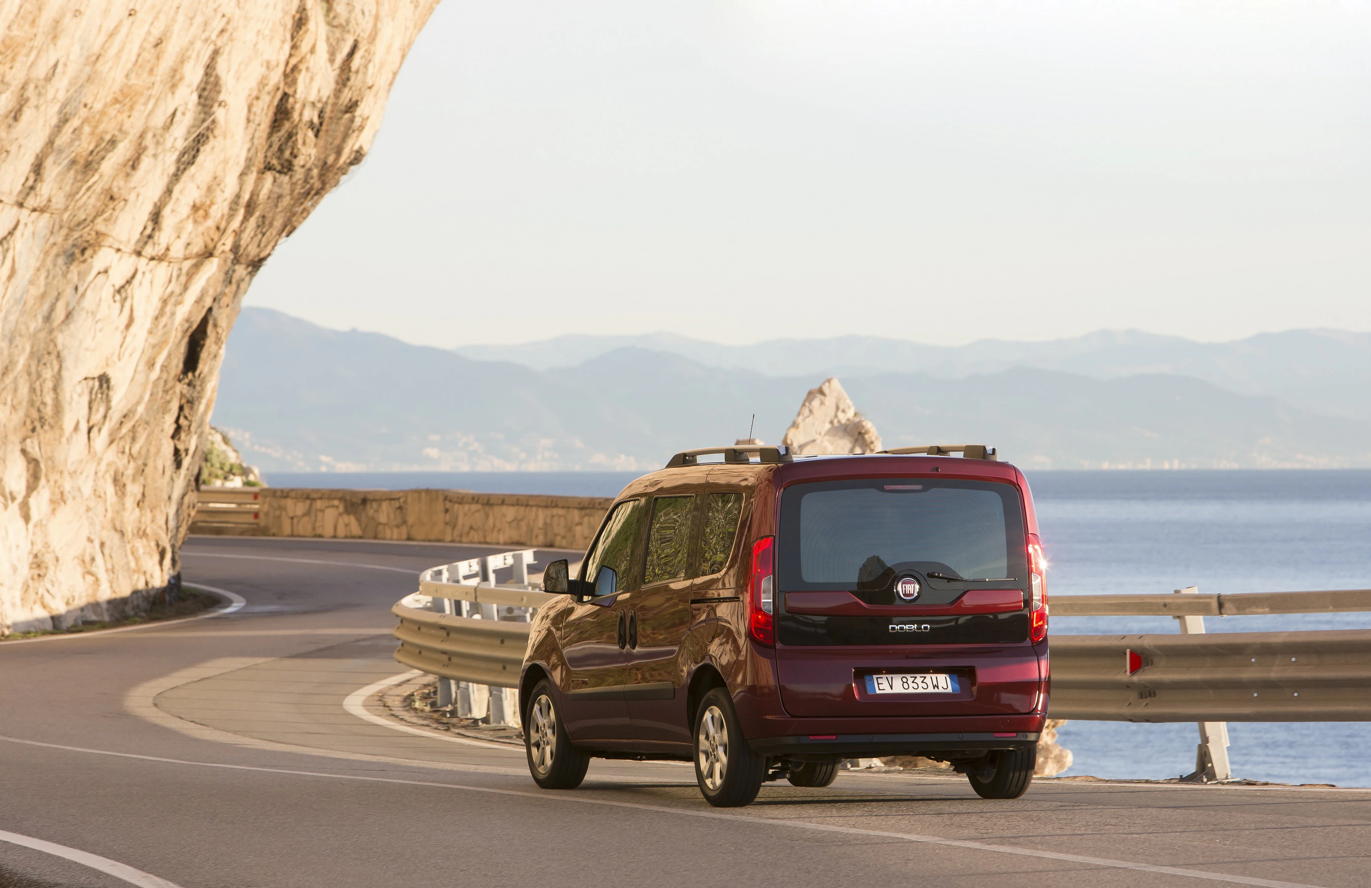 Ein roter Fiat Doblo fährt auf einer kurvigen Küstenstraße entlang eines felsigen Abhangs mit Blick auf das Meer und Berge im Hintergrund.