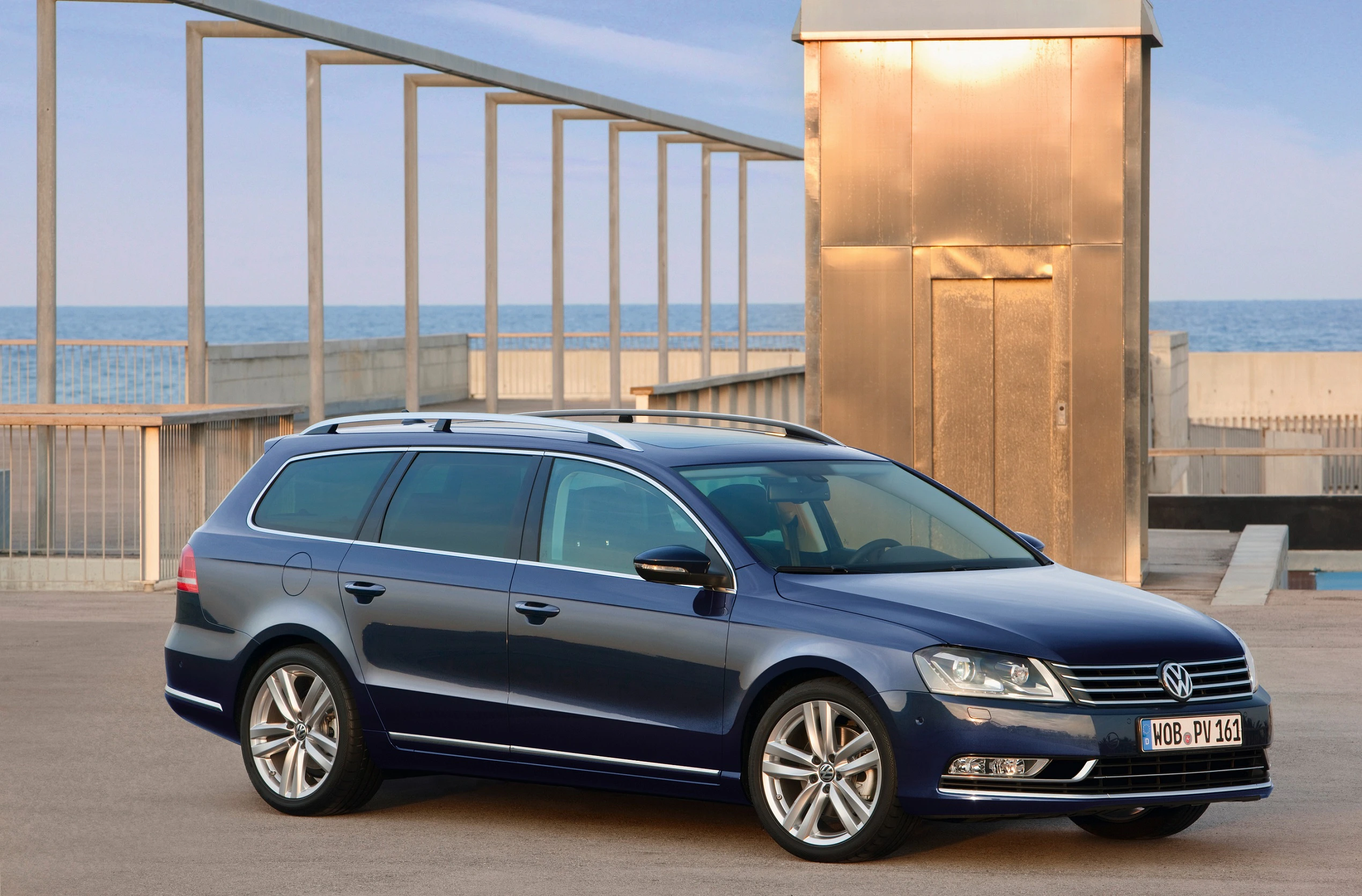 Blauer VW Passat Kombi parkt auf leerem Gelände vor moderner Architektur, im Hintergrund Meer und Himmel.