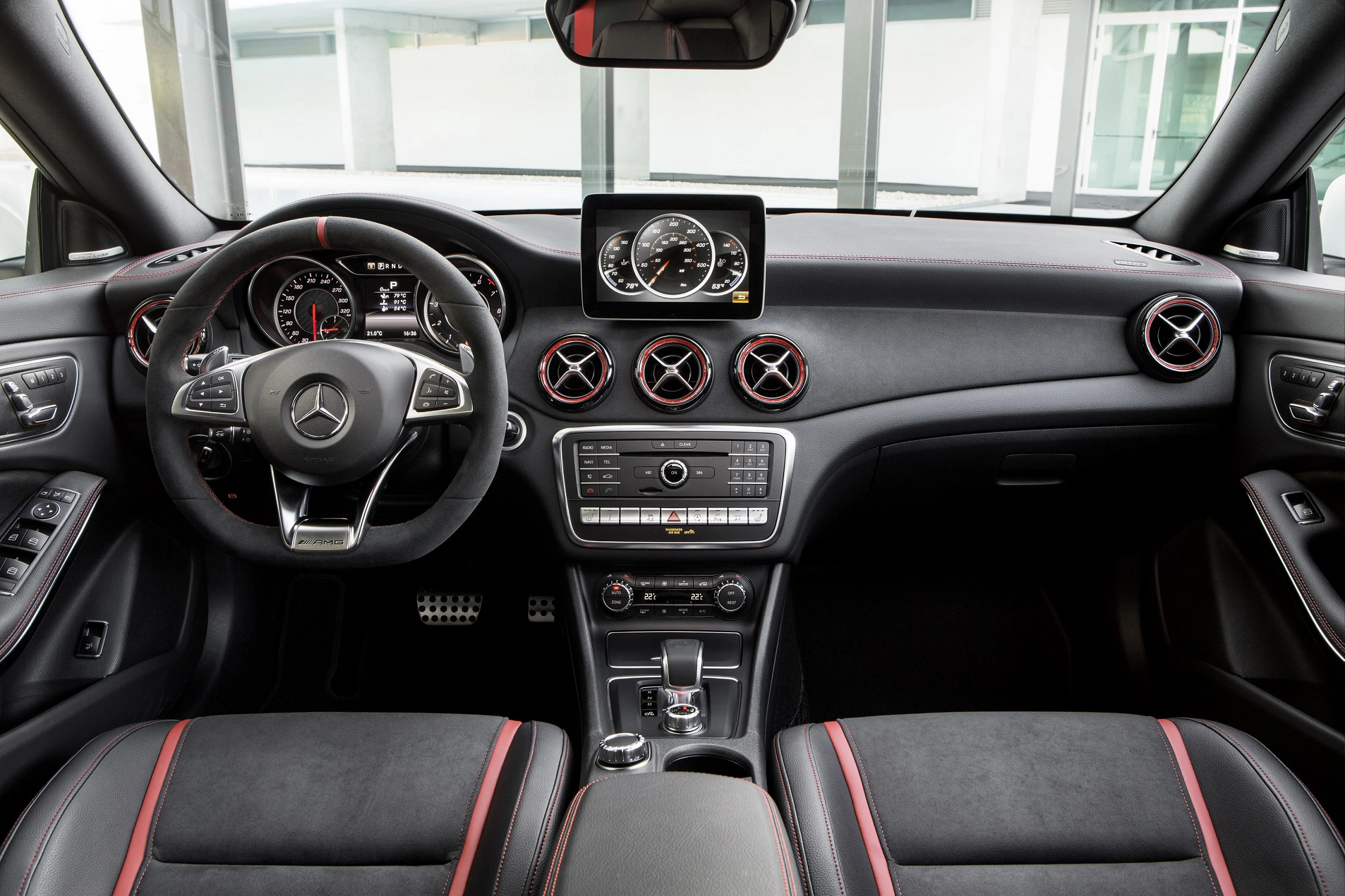 Innenraum eines Autos mit Mercedes-Logo, zentralem Bildschirm, Lüftungsdüsen in Rot-Schwarz, sportlichem Lenkrad und schwarzen Sitzen mit roten Akzenten.