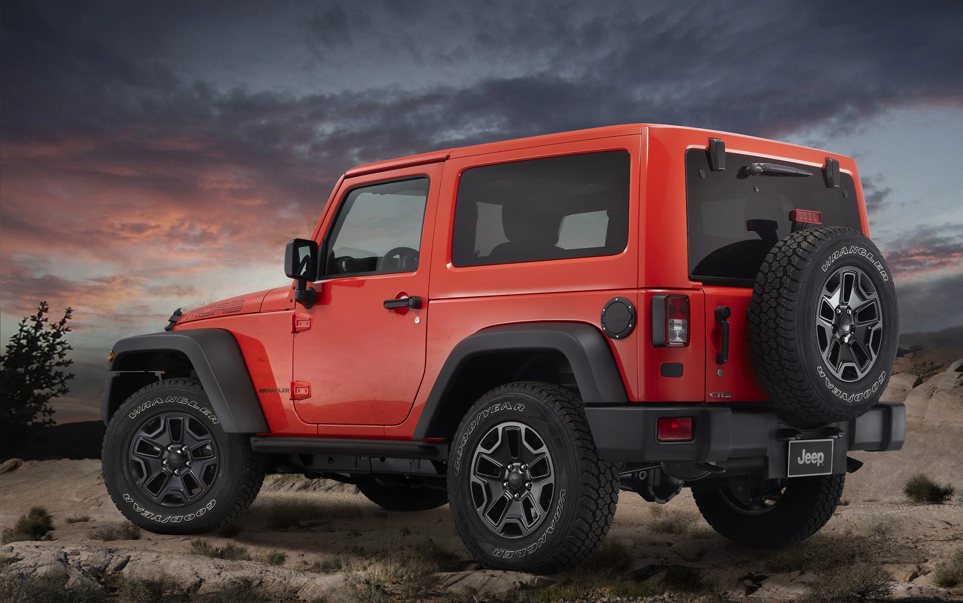Ein roter Geländewagen vom Typ Jeep steht in einer Wüstenlandschaft bei Sonnenuntergang, mit Reserverad hinten und markanten Reifen.