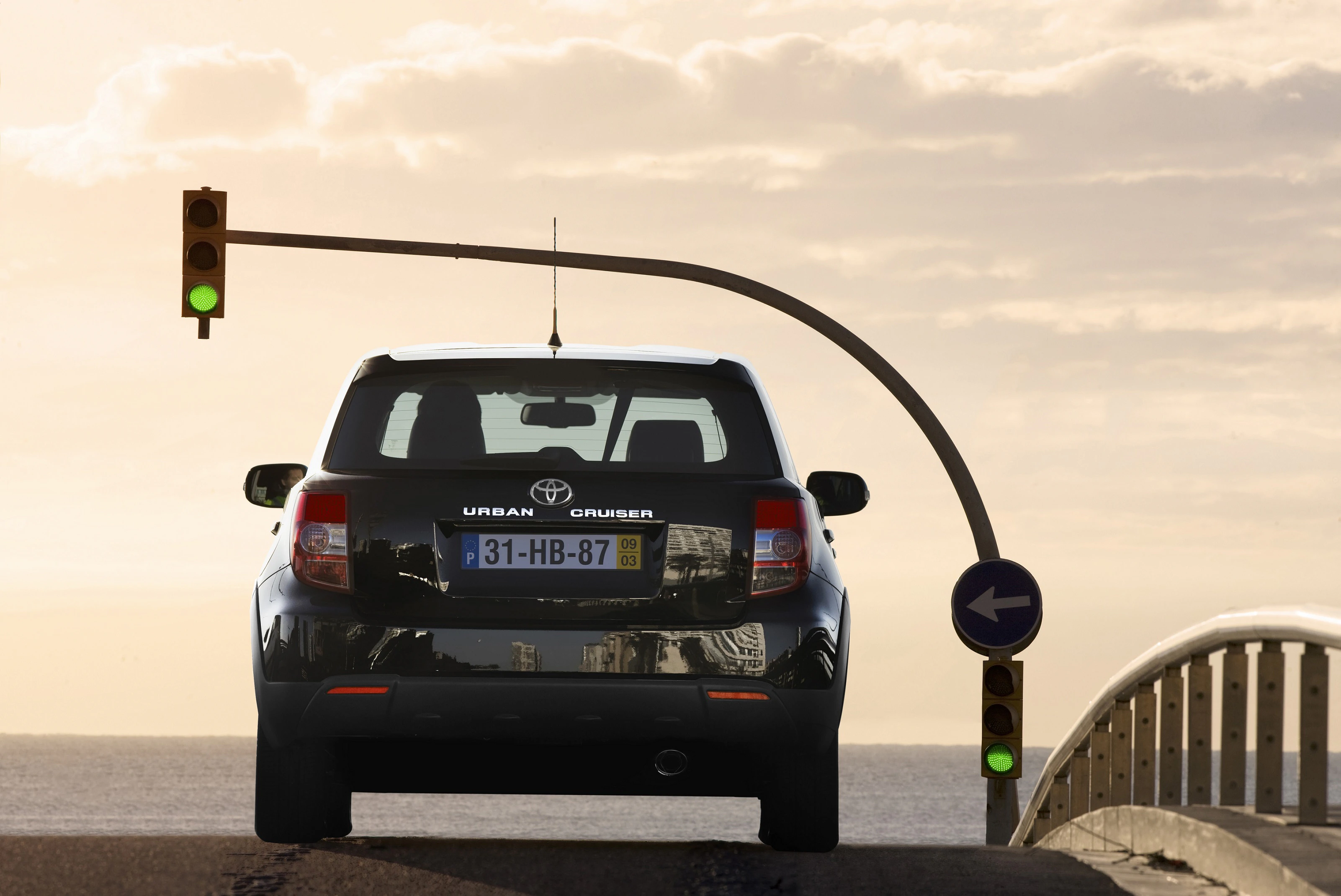 Auto an grüner Ampel auf einer Brücke, fährt Richtung Sonnenuntergang. Wolken und Meer im Hintergrund.