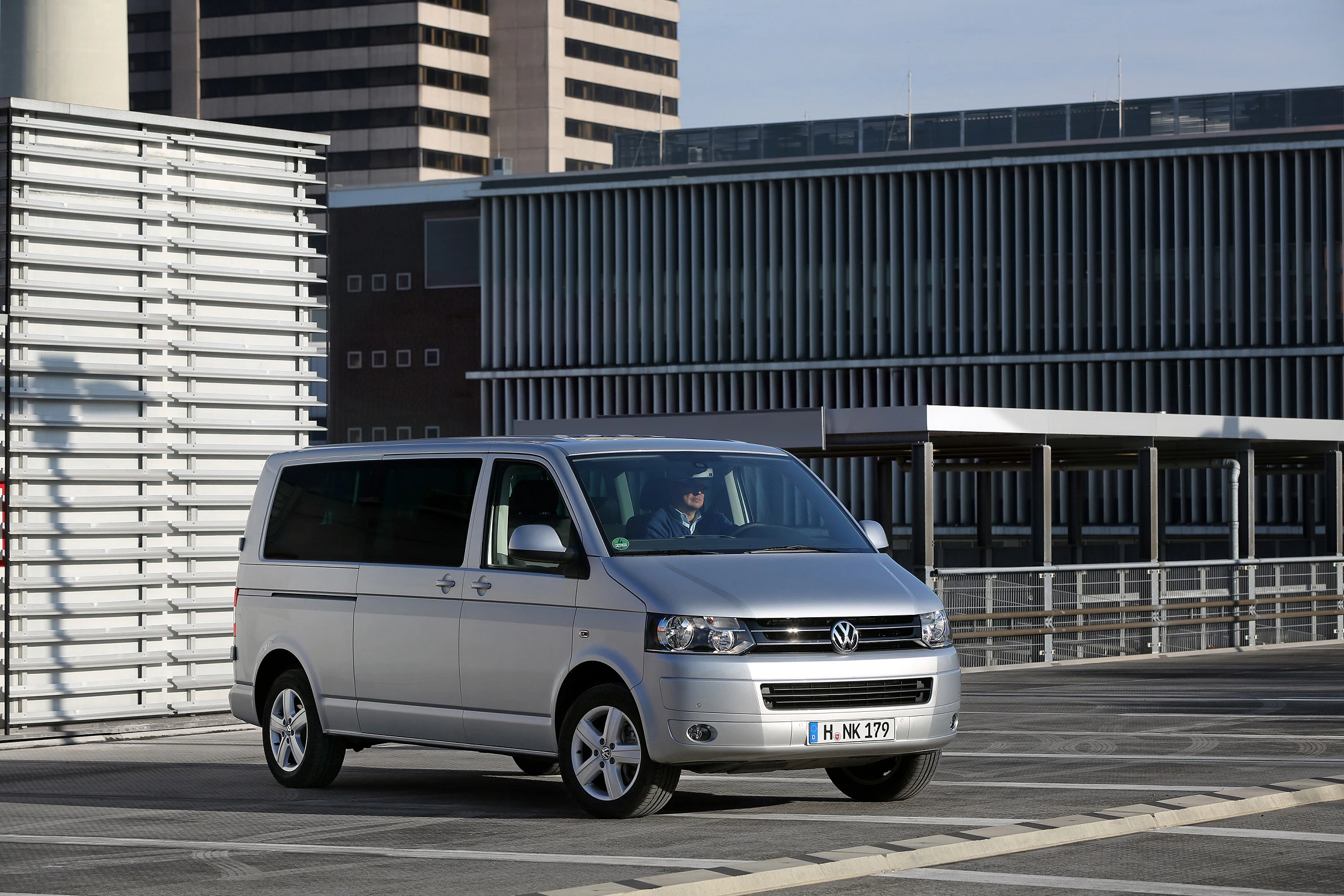Silberner Volkswagen-Van parkt auf einem modernen Parkdeck mit Bürogebäuden im Hintergrund an einem sonnigen Tag.