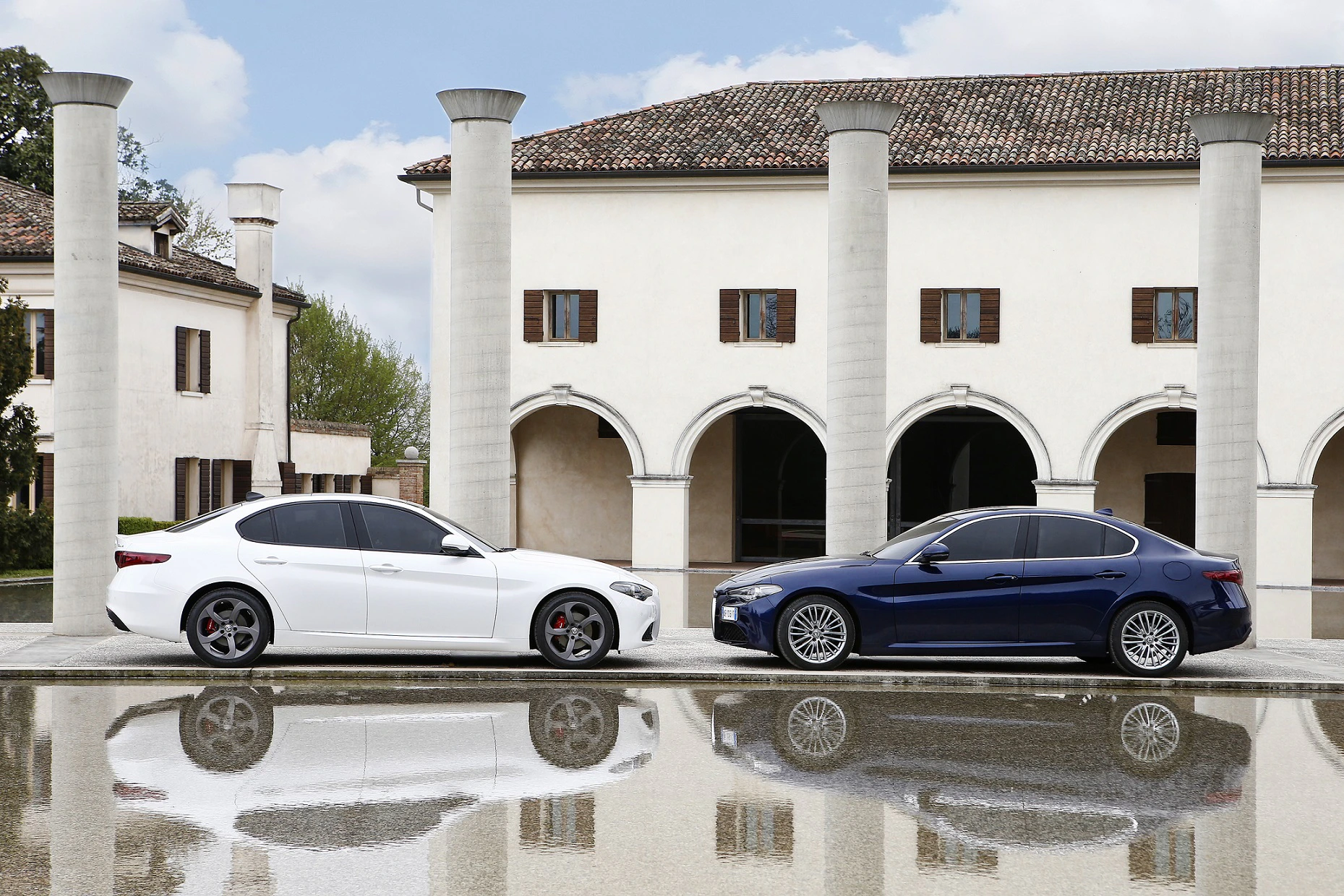 Zwei Autos, eines weiß und eines blau, stehen gegenüber in einer eleganten Hofkulisse mit Säulen, die sich im Wasser spiegeln.