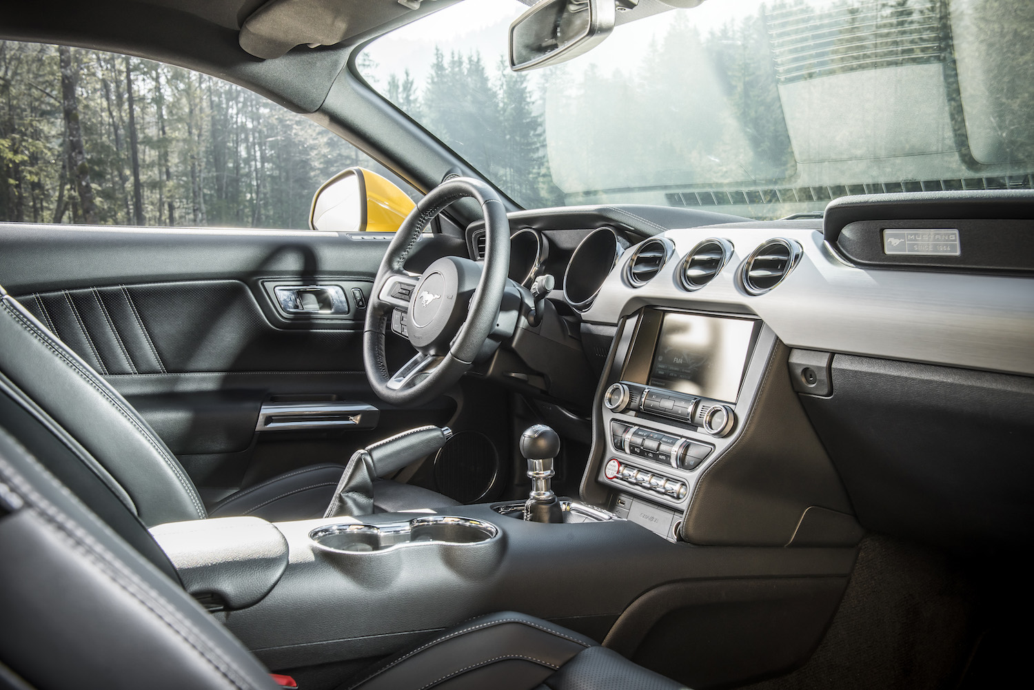 Innenraum eines modernen Sportwagens mit Lederpolstern, sportlichem Lenkrad, Schalthebel und digitalem Display; Blick auf bewaldete Umgebung.