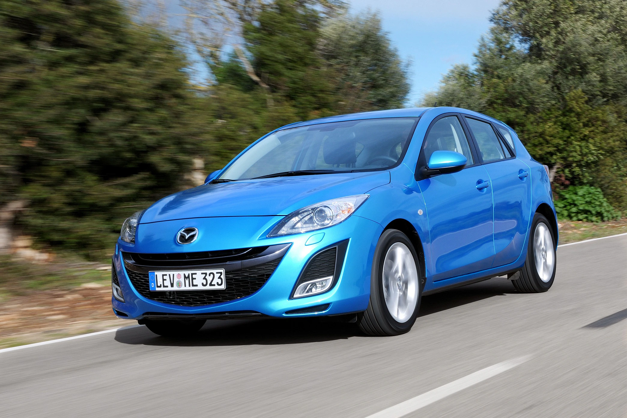 Blauer Mazda auf einer Landstraße, umgeben von grünen Bäumen und blauem Himmel, fährt dynamisch nach links.