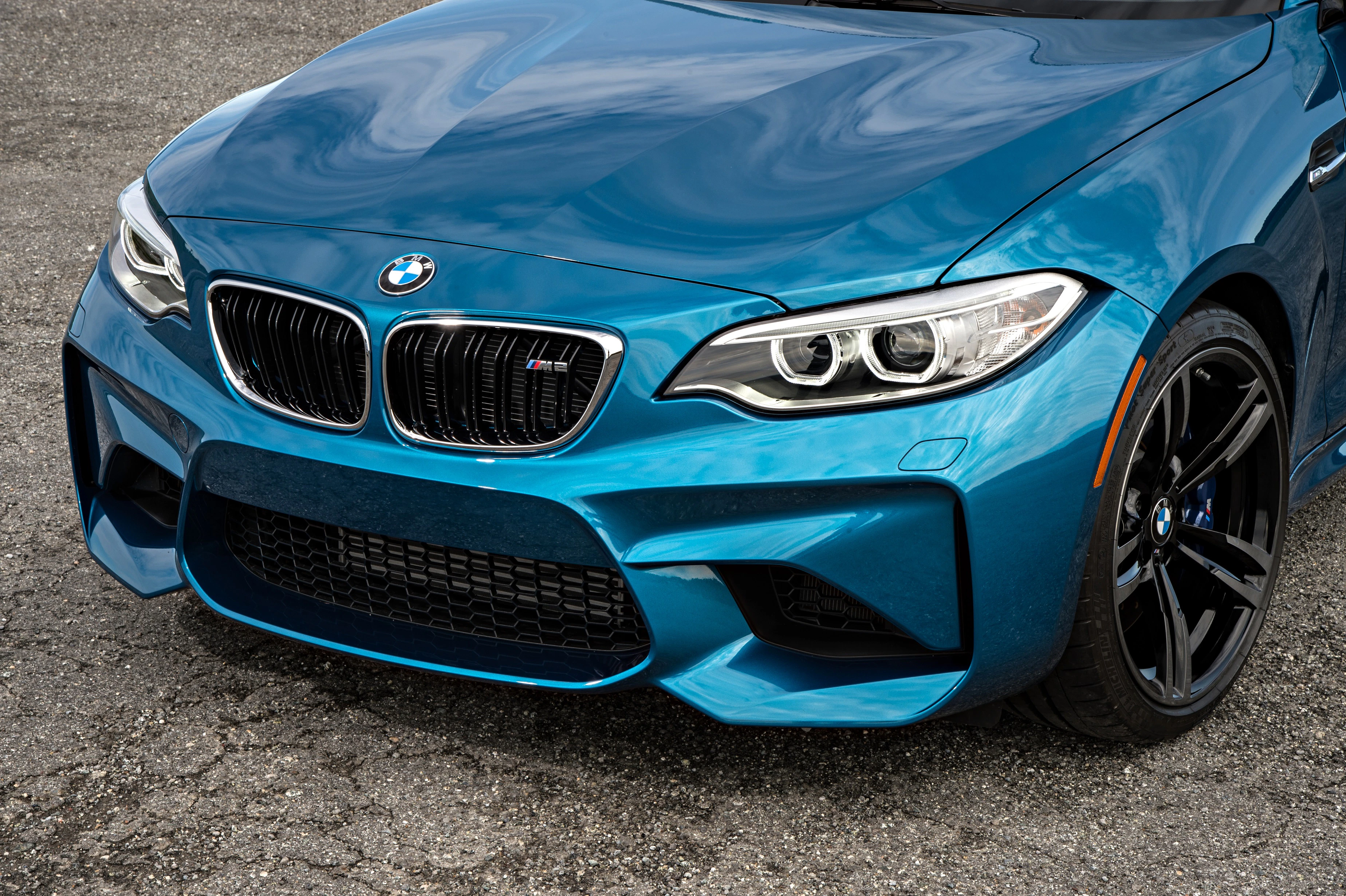 Frontansicht eines blauen BMW M2 mit markanten Scheinwerfern und sportlicher Frontstoßstange auf grauem Asphalt.