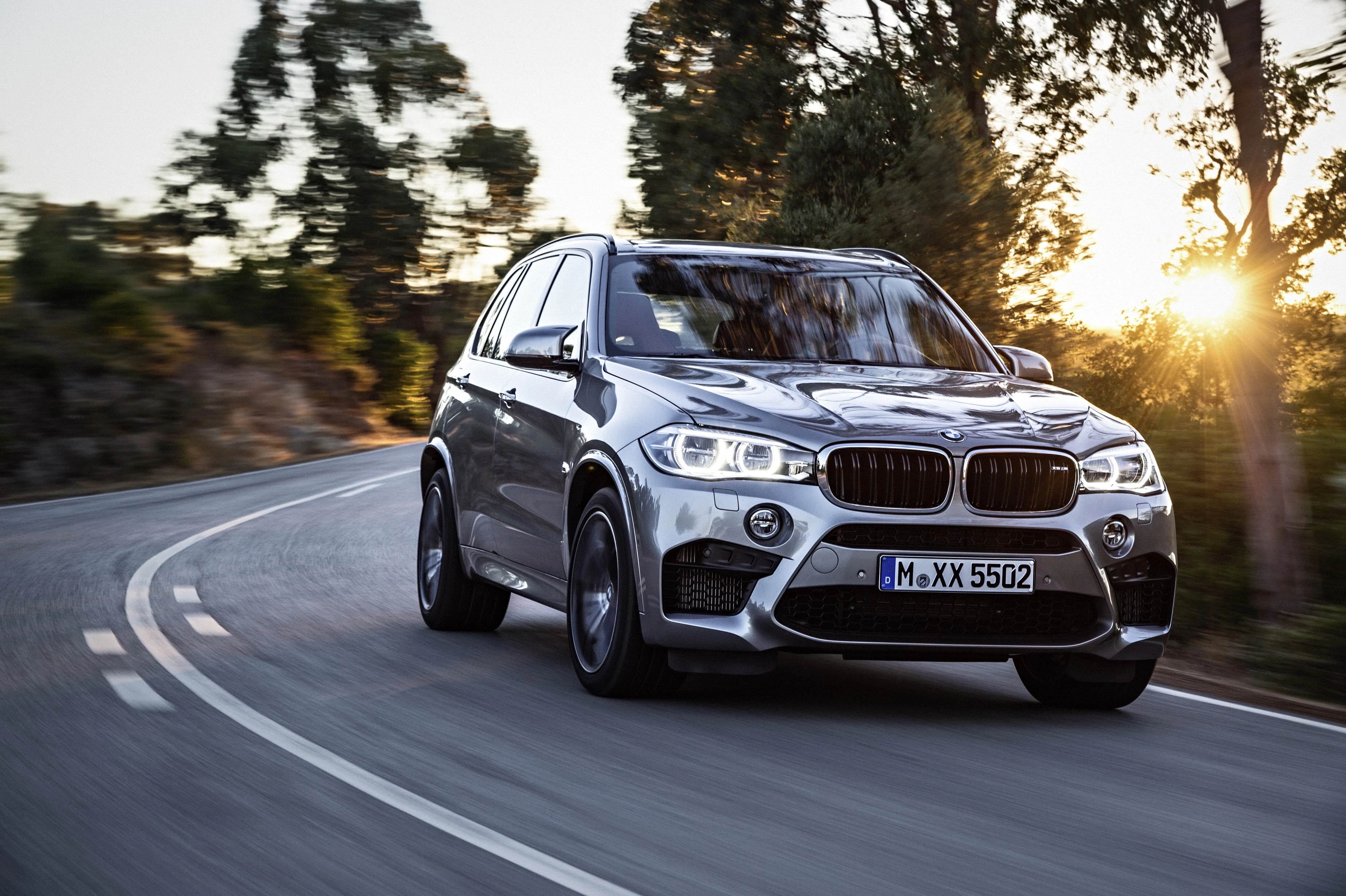 Silberner SUV fährt auf einer kurvigen Straße bei Sonnenuntergang, umgeben von Bäumen. Vorne ist die markante BMW-Niere erkennbar.