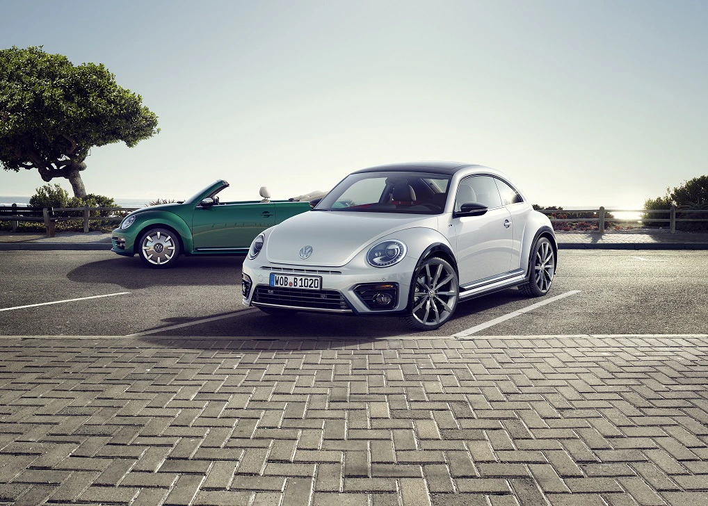 Zwei VW Beetle auf einem Parkplatz nahe der Küste, einer weiß geschlossen, der andere grün als Cabrio. Im Hintergrund Meer und Bäume.