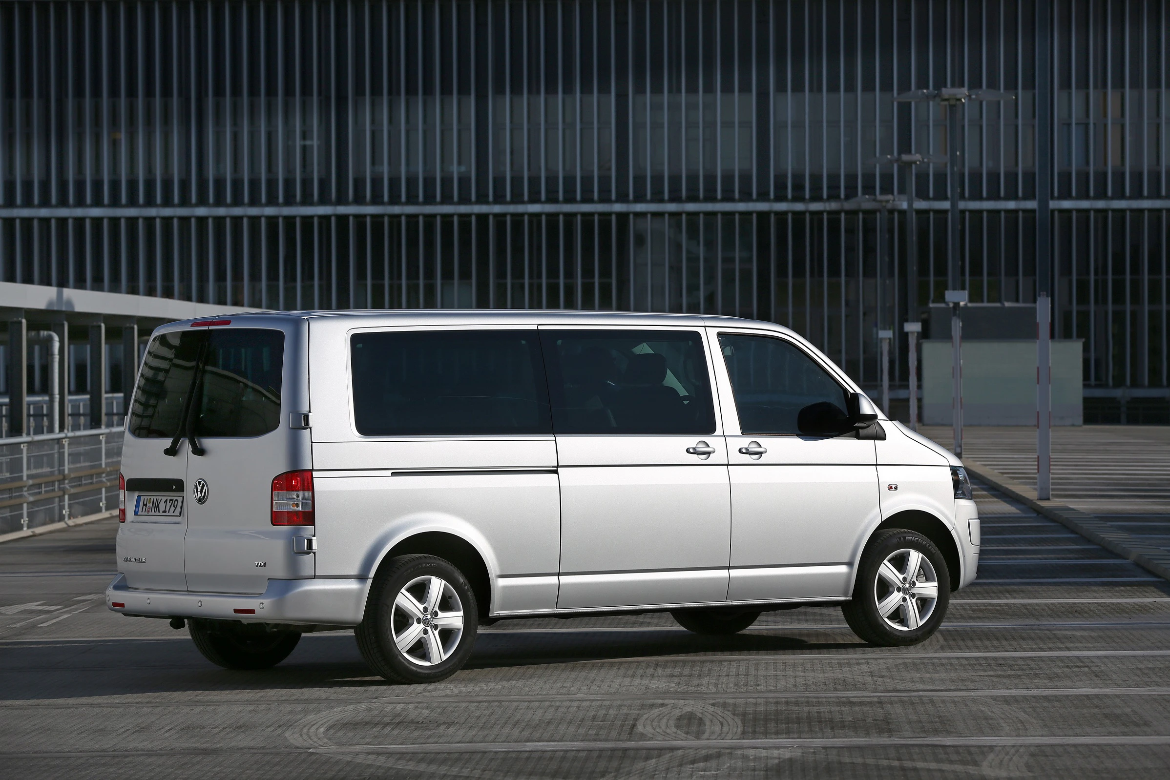 Silberner VW-Transporter parkt auf einem leeren Außengelände vor einem modernen Gebäude.