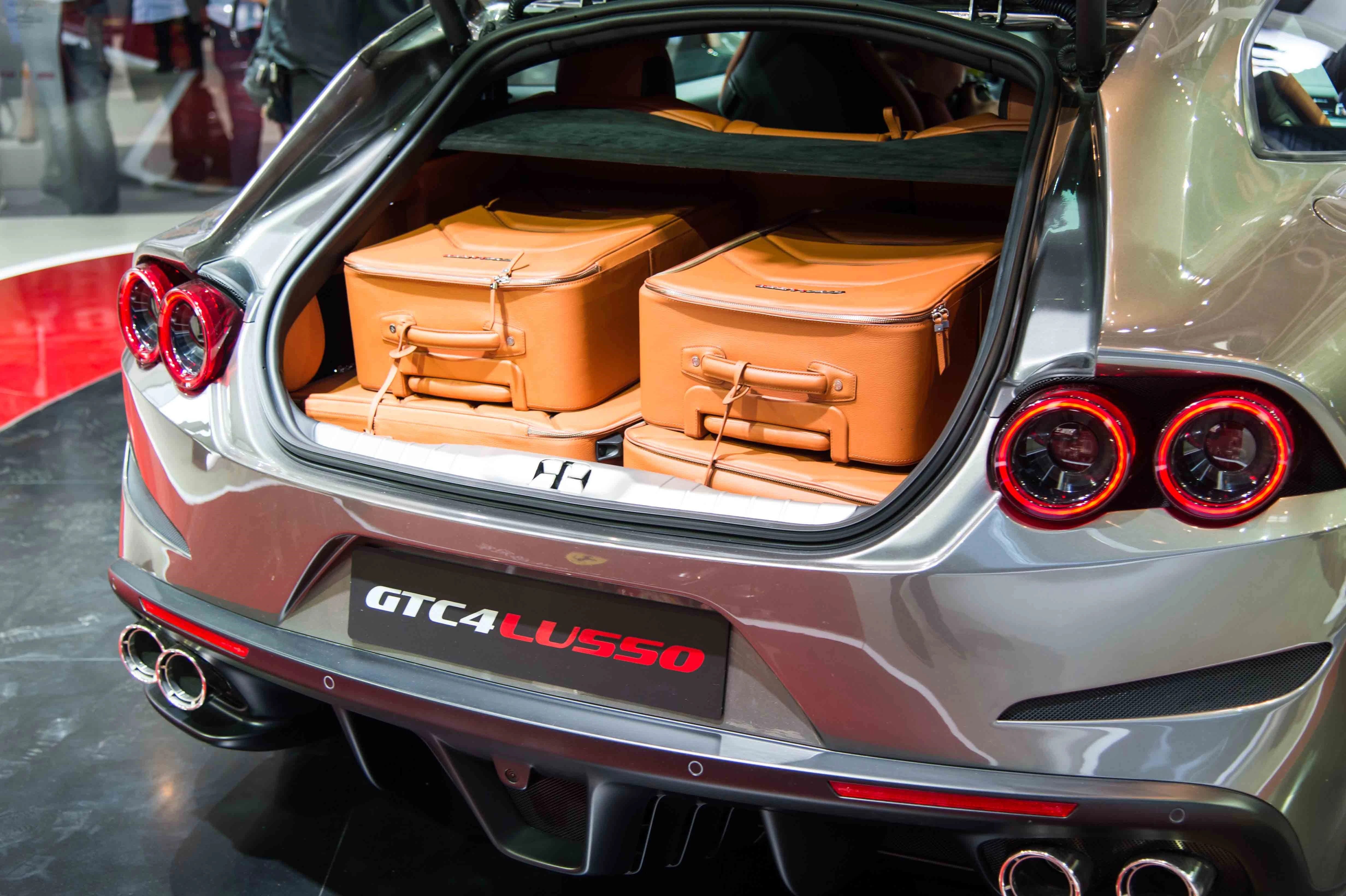 Silbernes Auto mit geöffnetem Kofferraum, darin zwei braune Reisetaschen. Rücklichter und Auspuff sichtbar, Modell GTC4Lusso.