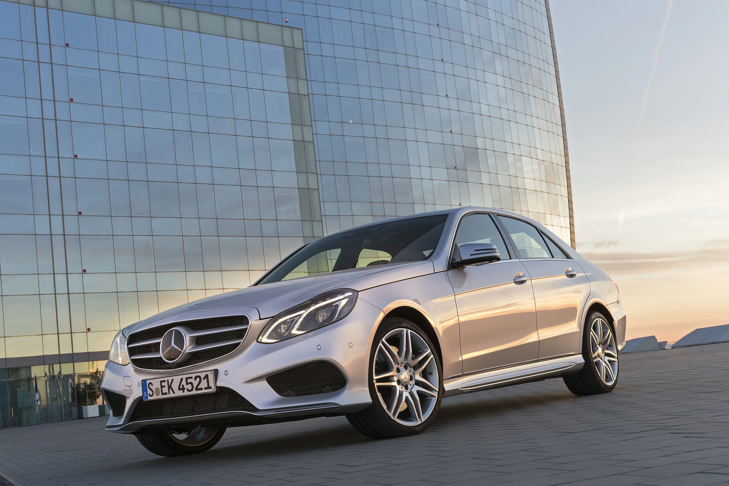 Silberner Mercedes-Benz steht vor einem modernen Glasgebäude bei Sonnenuntergang. Elegantes Design und stilvolle Architektur.