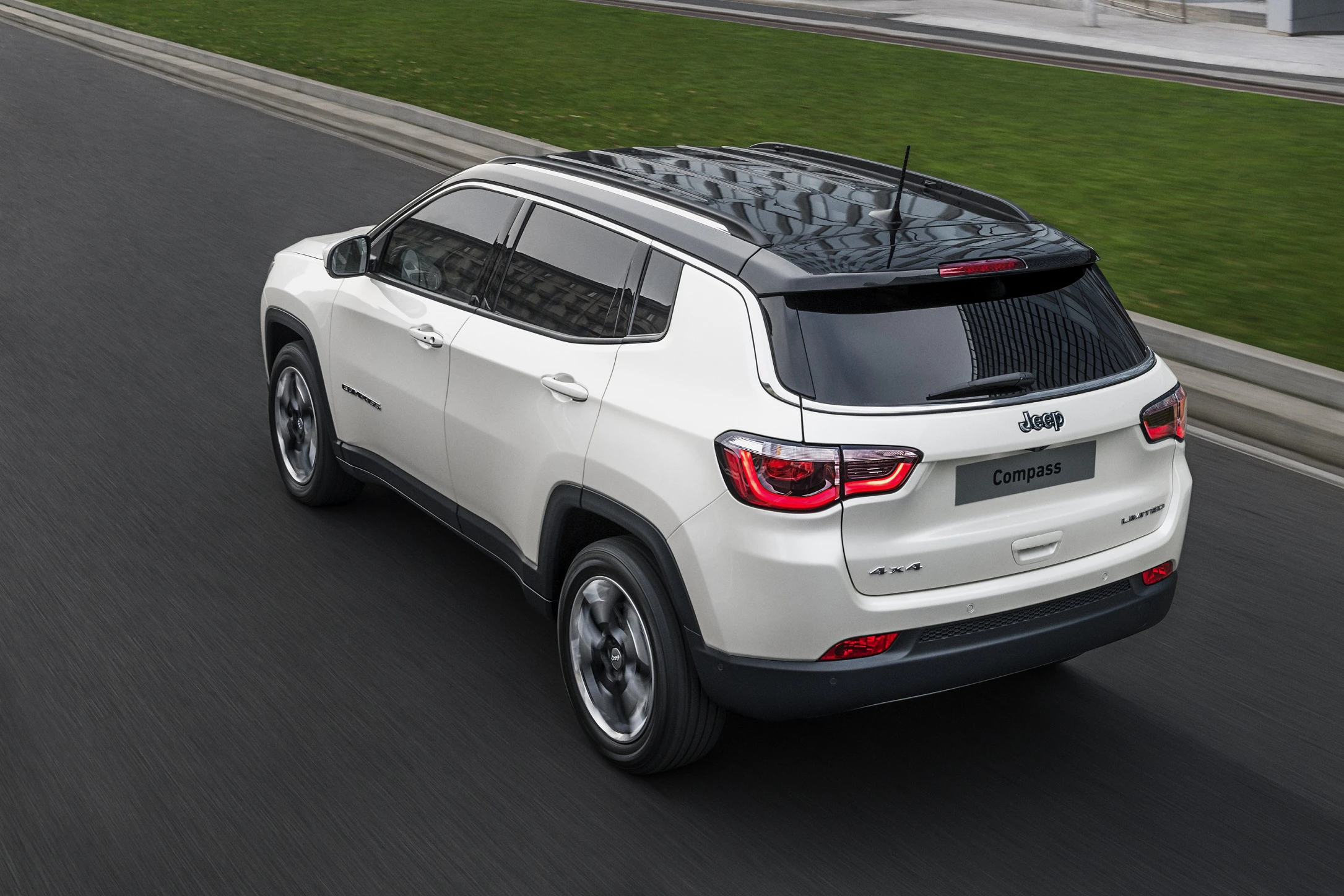 Weißer Jeep Compass SUV fährt auf einer Asphaltstraße, vorbei an grünem Rasen und modernen Gebäude; Ansicht von hinten oben.