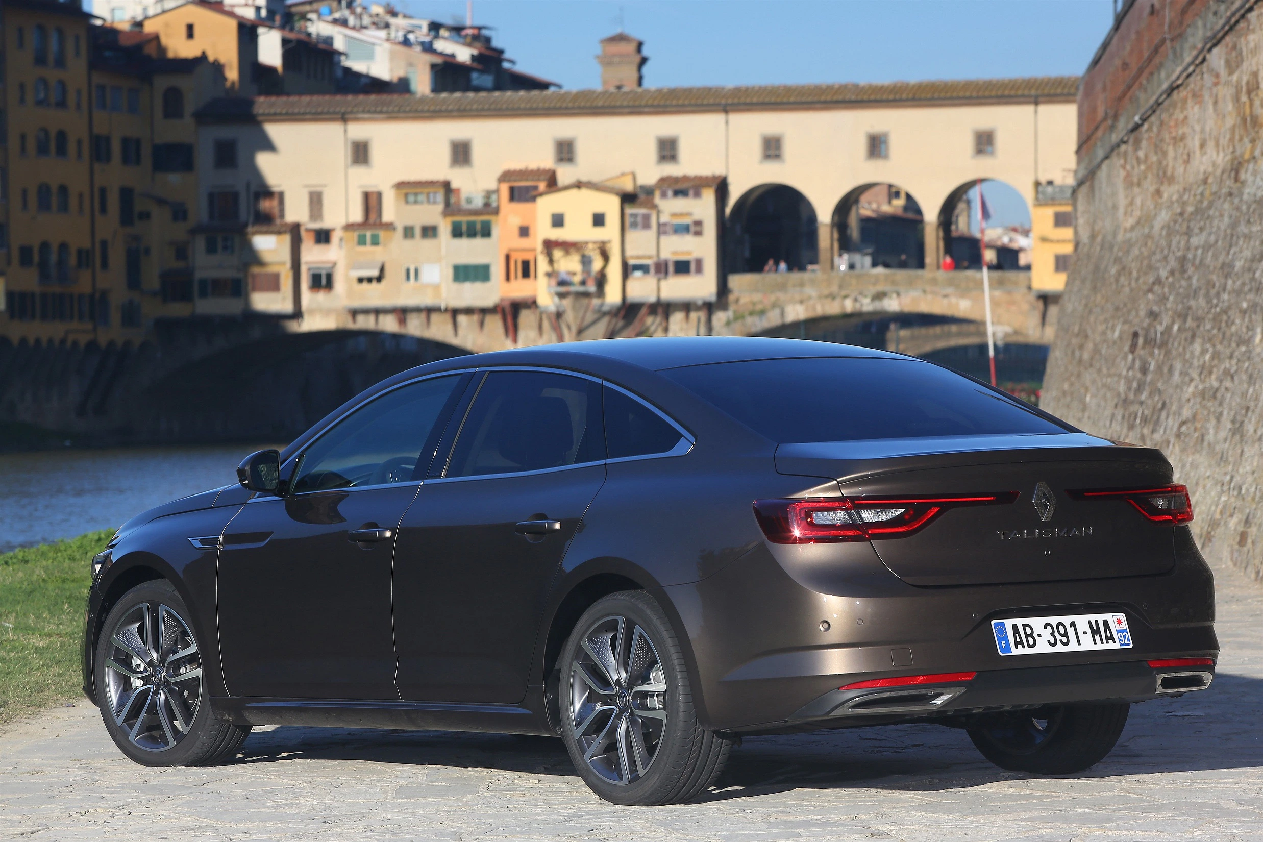 Brauner Renault Talisman parkt vor einer historischen Brücke in einer Stadt bei sonnigem Wetter.