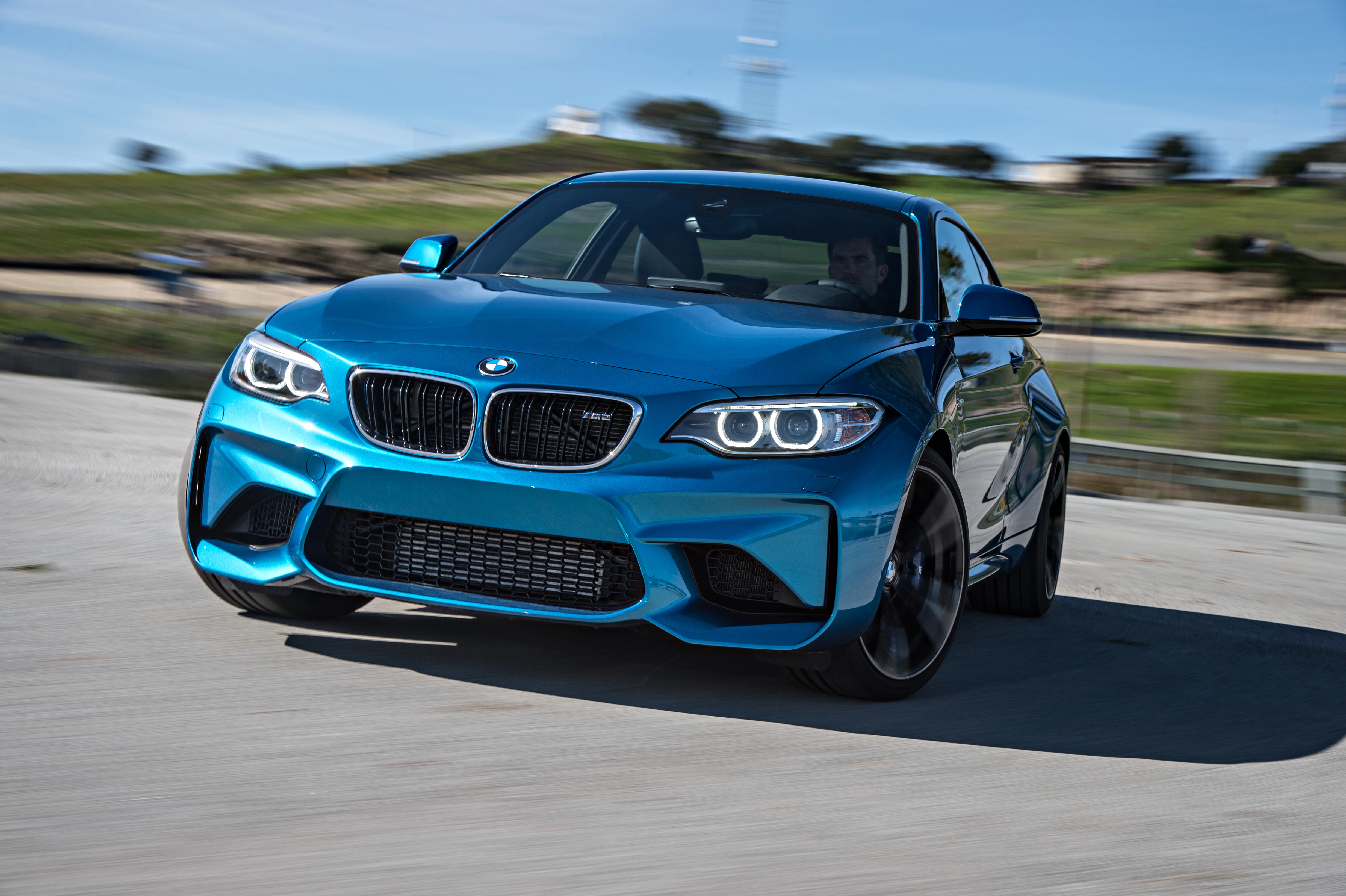 Ein blauer BMW M2 fährt bei sonnigem Wetter auf einer Landstraße, Hintergrund zeigt grüne Hügel und blauen Himmel.