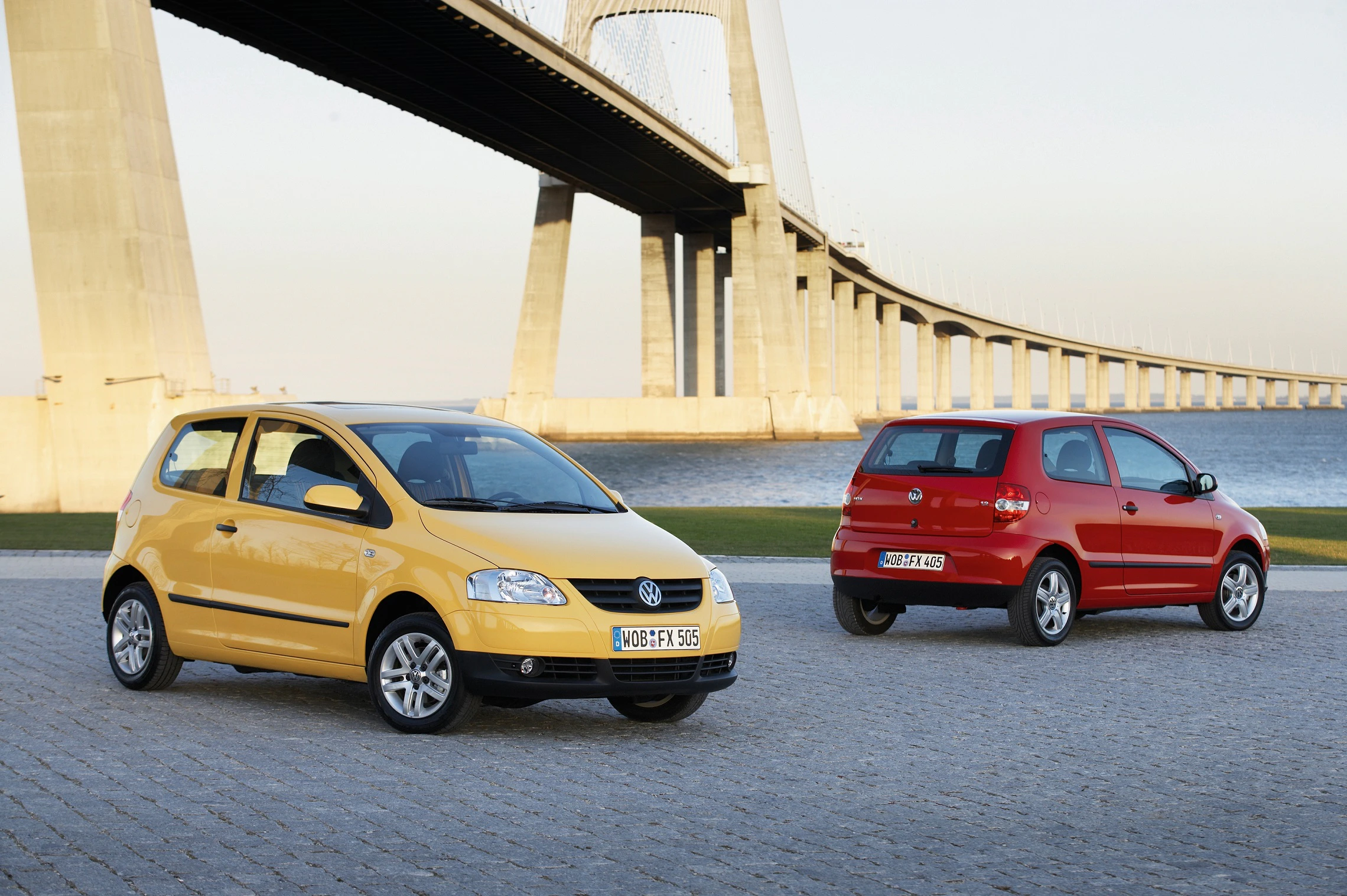 Zwei VW-Fox-Autos, eines gelb und eines rot, stehen auf einer gepflasterten Fläche vor einer großen Brücke bei Tageslicht.