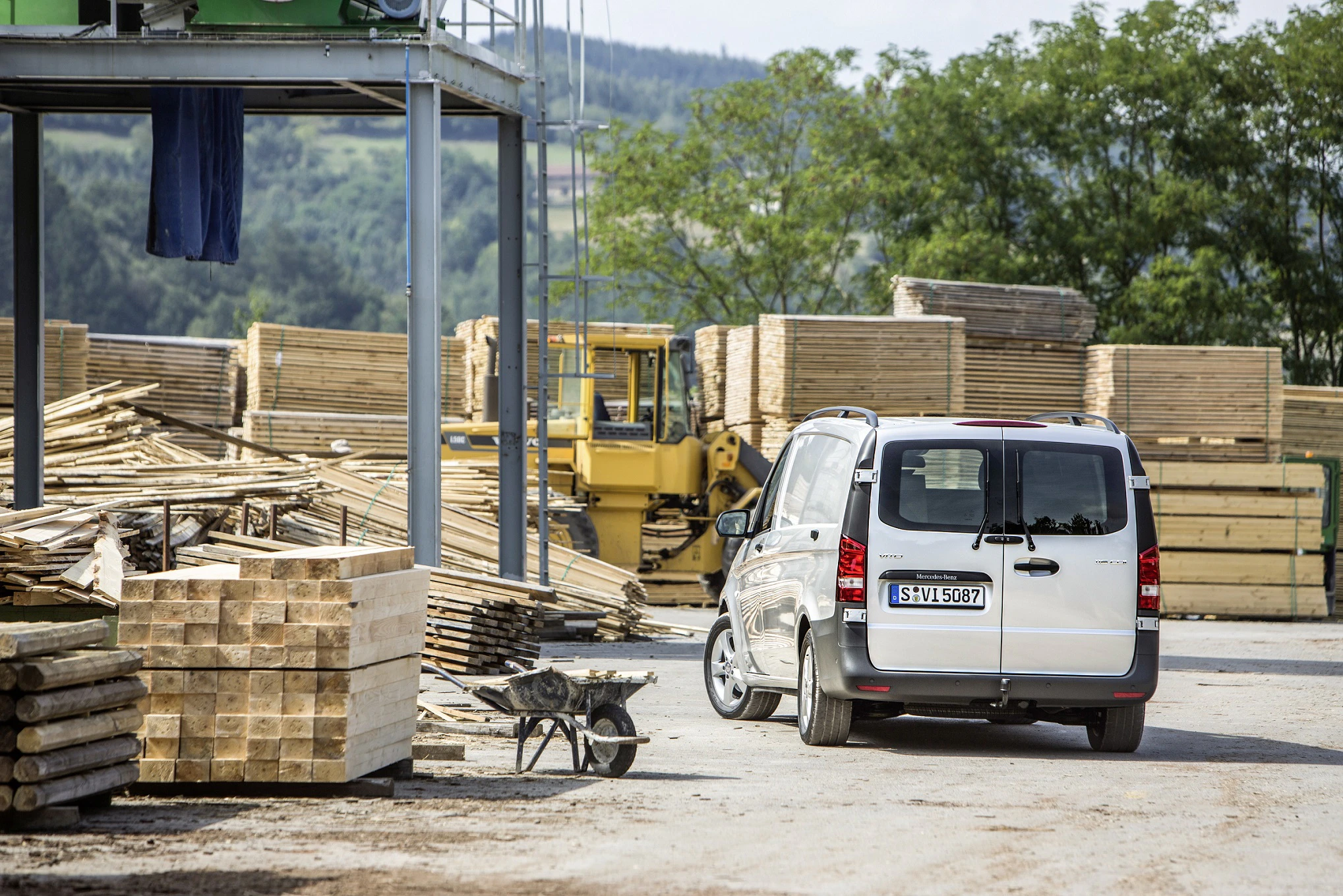 Ein weißer Lieferwagen parkt auf einem Holzplatz neben gestapelten Holzstämmen und einer Schubkarre, im Hintergrund ein gelber Radlader.