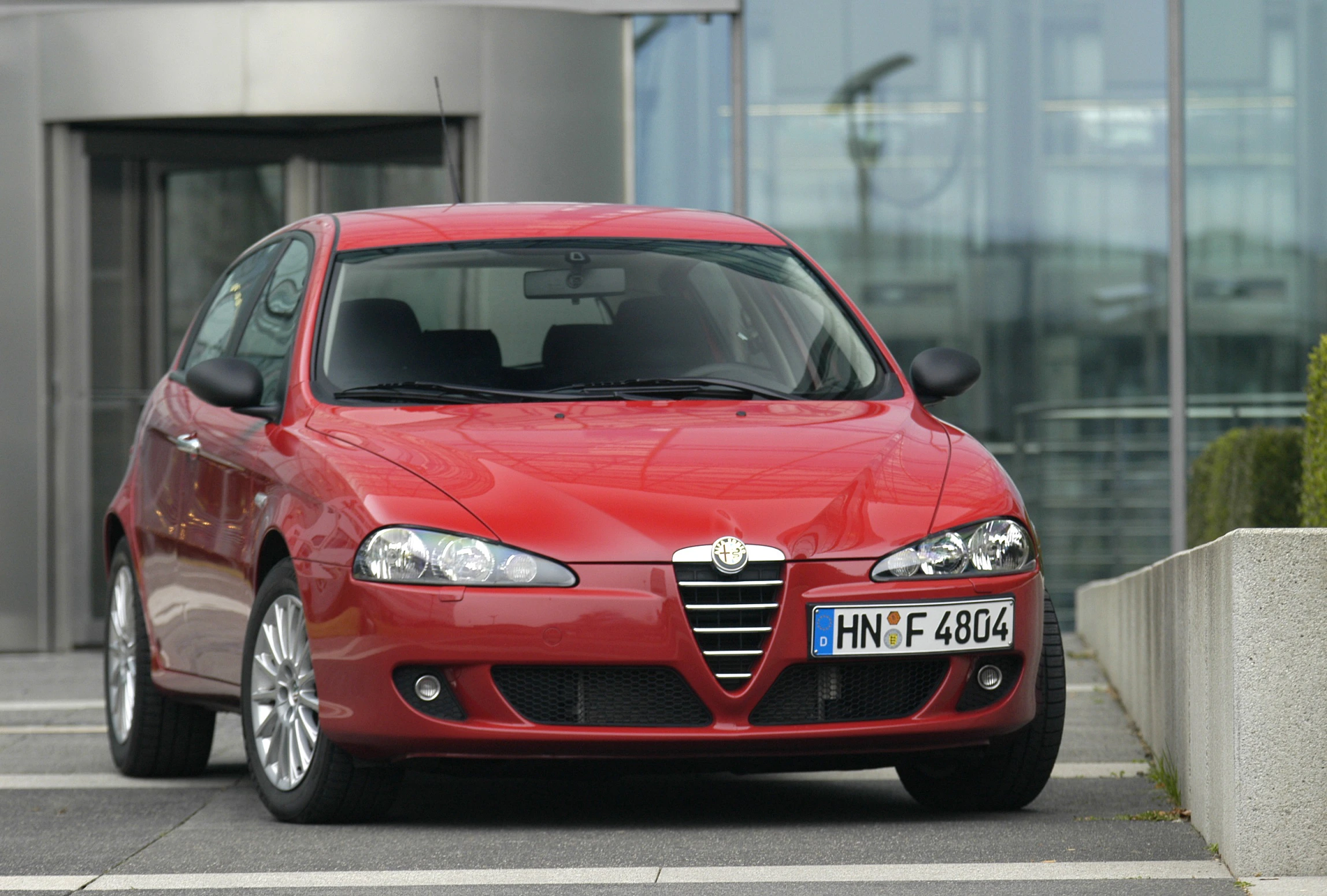 Roter Alfa Romeo 147 steht vor einem modernen Gebäude. Das Auto hat deutsche Nummernschilder und ist auf einem Parkplatz abgestellt.