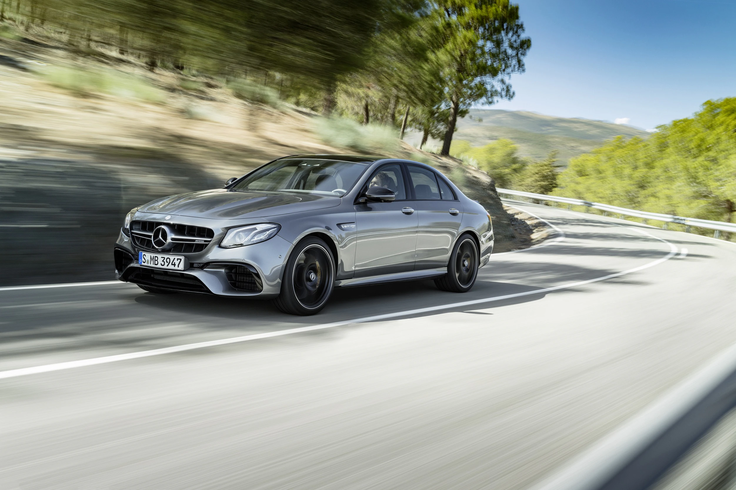 Silberfarbener Mercedes auf kurviger Bergstraße, umgeben von Bäumen. Dynamische Fahrt bei Tageslicht.
