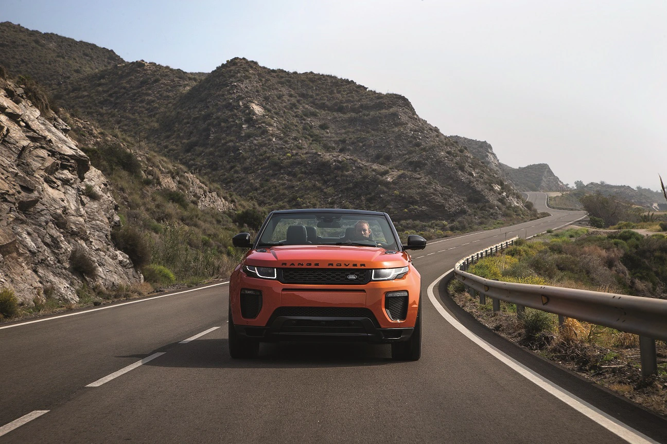 Ein orangefarbener SUV fährt auf einer kurvigen Straße durch eine bergige Landschaft unter klarem Himmel.