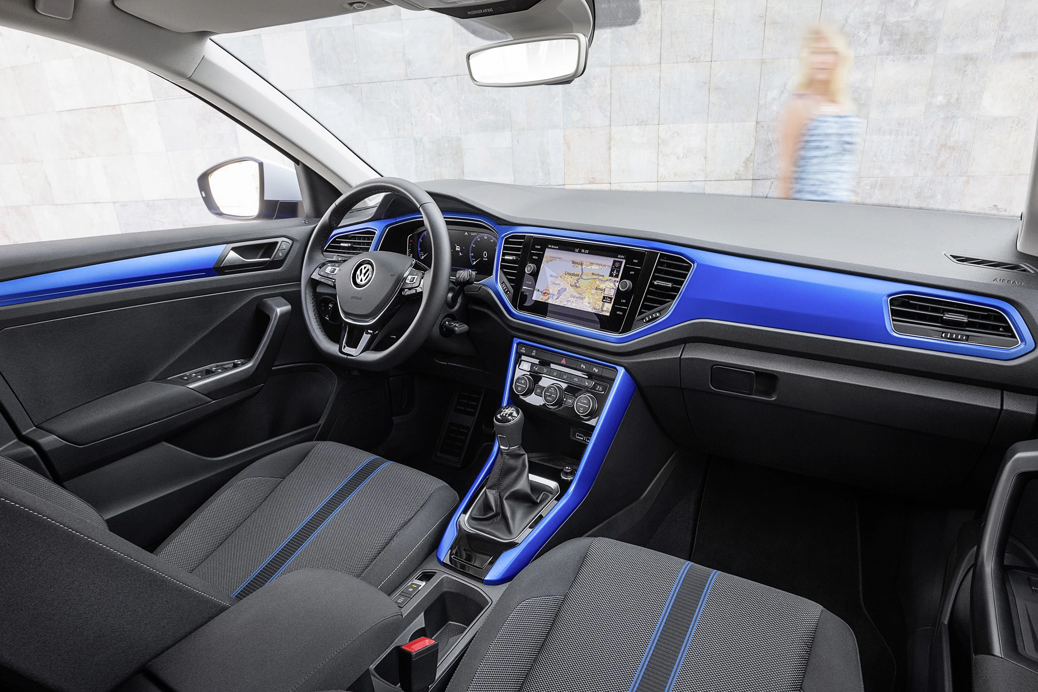 Innenraum eines modernen Autos mit blauen Akzenten, Lenkrad und Touchscreen-Display; draußen unscharfe Person im Hintergrund.