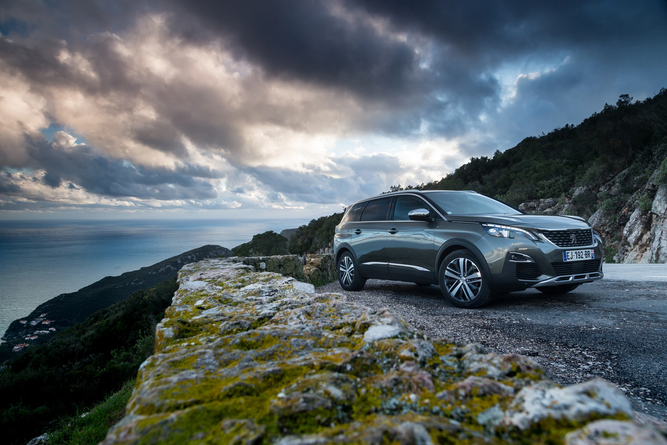 SUV auf kurviger Küstenstraße, im Hintergrund dramatischer Himmel und Meer, umgeben von bewaldeten Hügeln und steiniger Mauer.