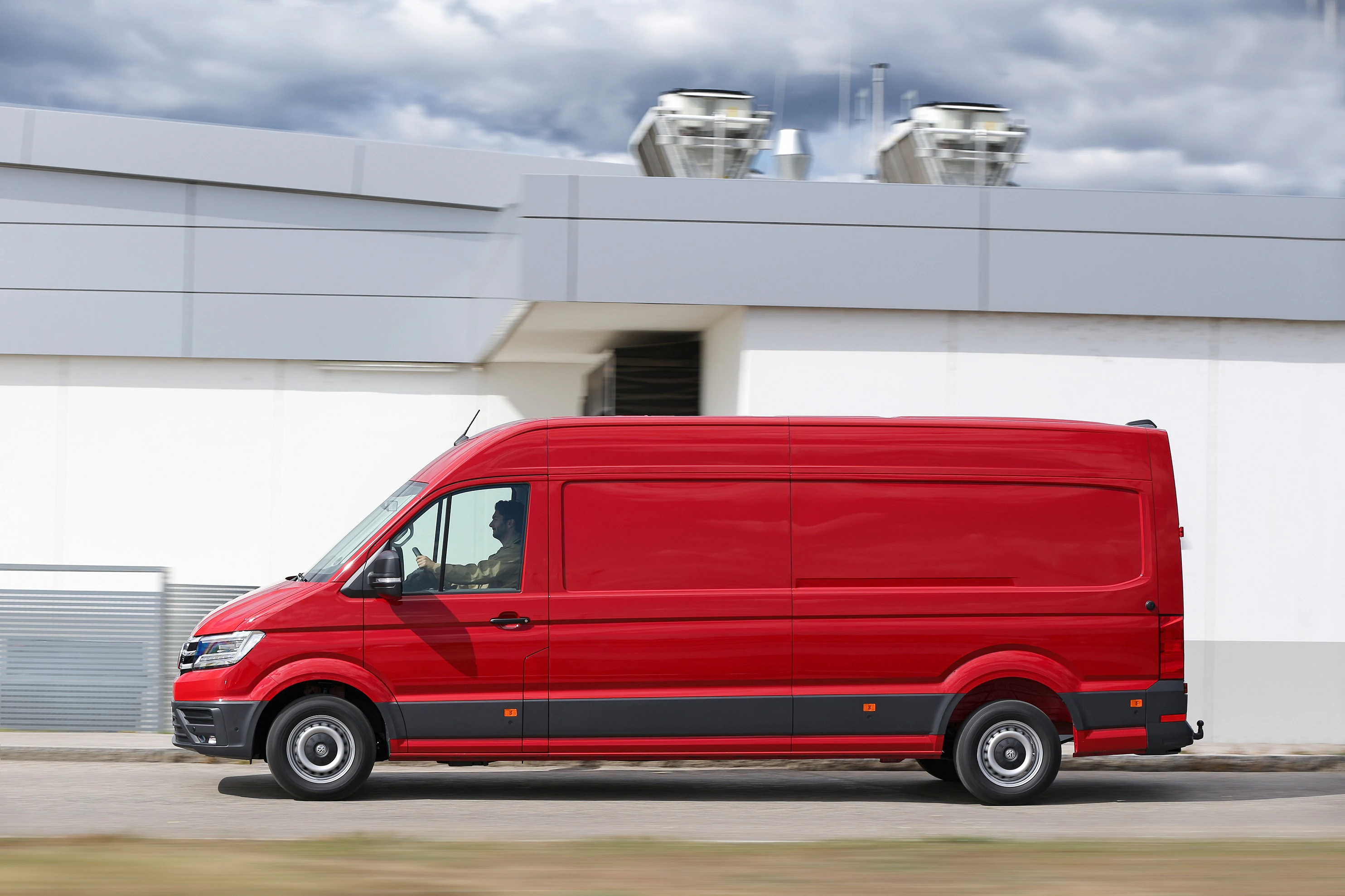 Roter Lieferwagen fährt entlang eines Industriegebäudes. Fahrer im Inneren, bewölkter Himmel im Hintergrund.