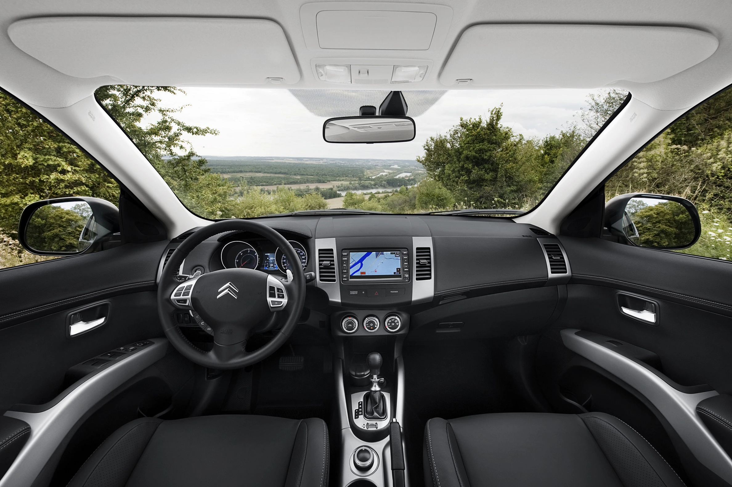 Innenansicht eines Autos mit schwarzem Interieur, Blick auf Lenkrad, Mittelkonsole mit Bildschirm und weite Landschaft durch die Frontscheibe.