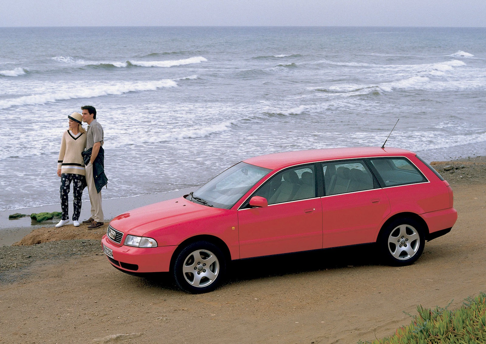 Roter Kombi am Strand, parkend mit einem Paar, das in die Ferne schaut, während die Wellen im Hintergrund an das Ufer schlagen.