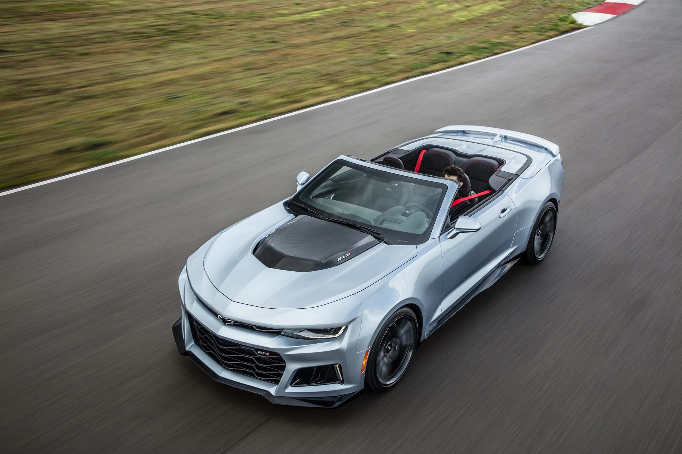 Silberner Cabrio-Sportwagen fährt auf einer Straße; dynamische Perspektive von oben, mit einem Fahrer am Steuer.