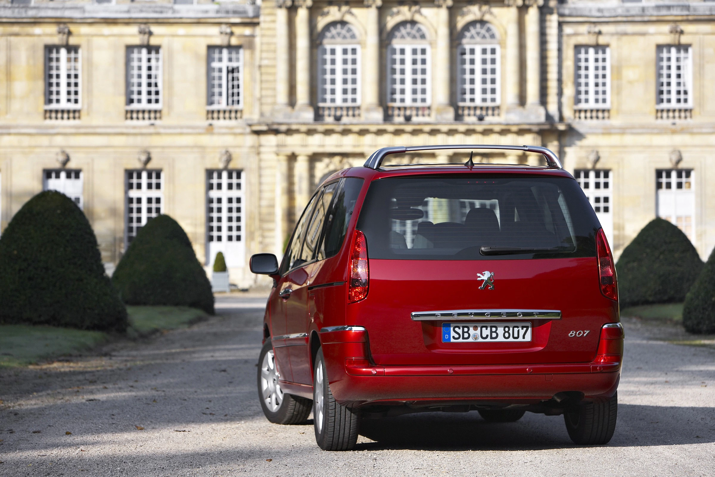 Roter Peugeot 807 parkt vor einem historischen Gebäude mit aufwendiger Fassade und gepflegten Büschen im Hintergrund.