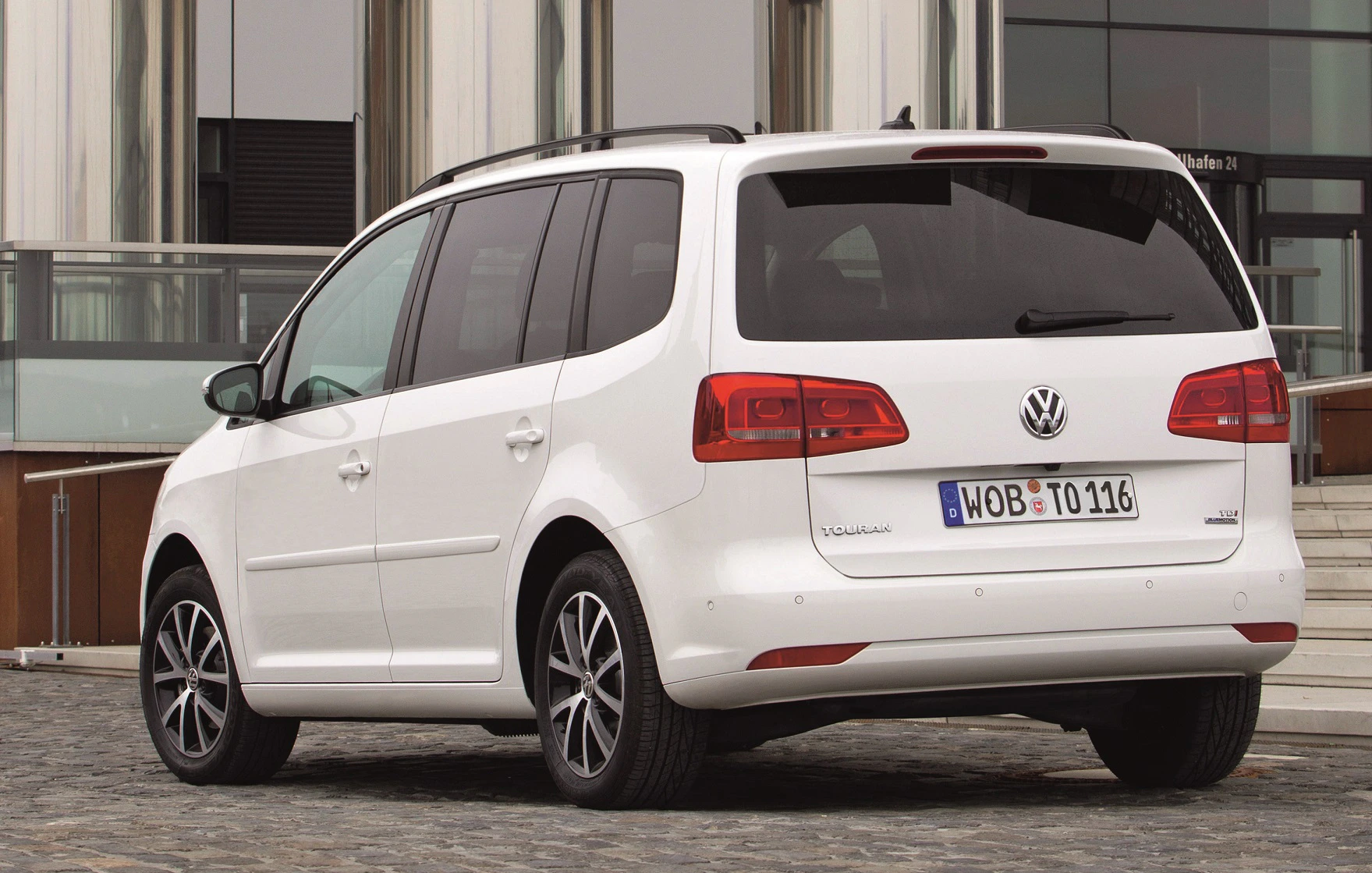 Weißer VW Touran parkt draußen vor einem modernen Gebäude. Rückansicht des Wagens mit schwarzen Felgen.