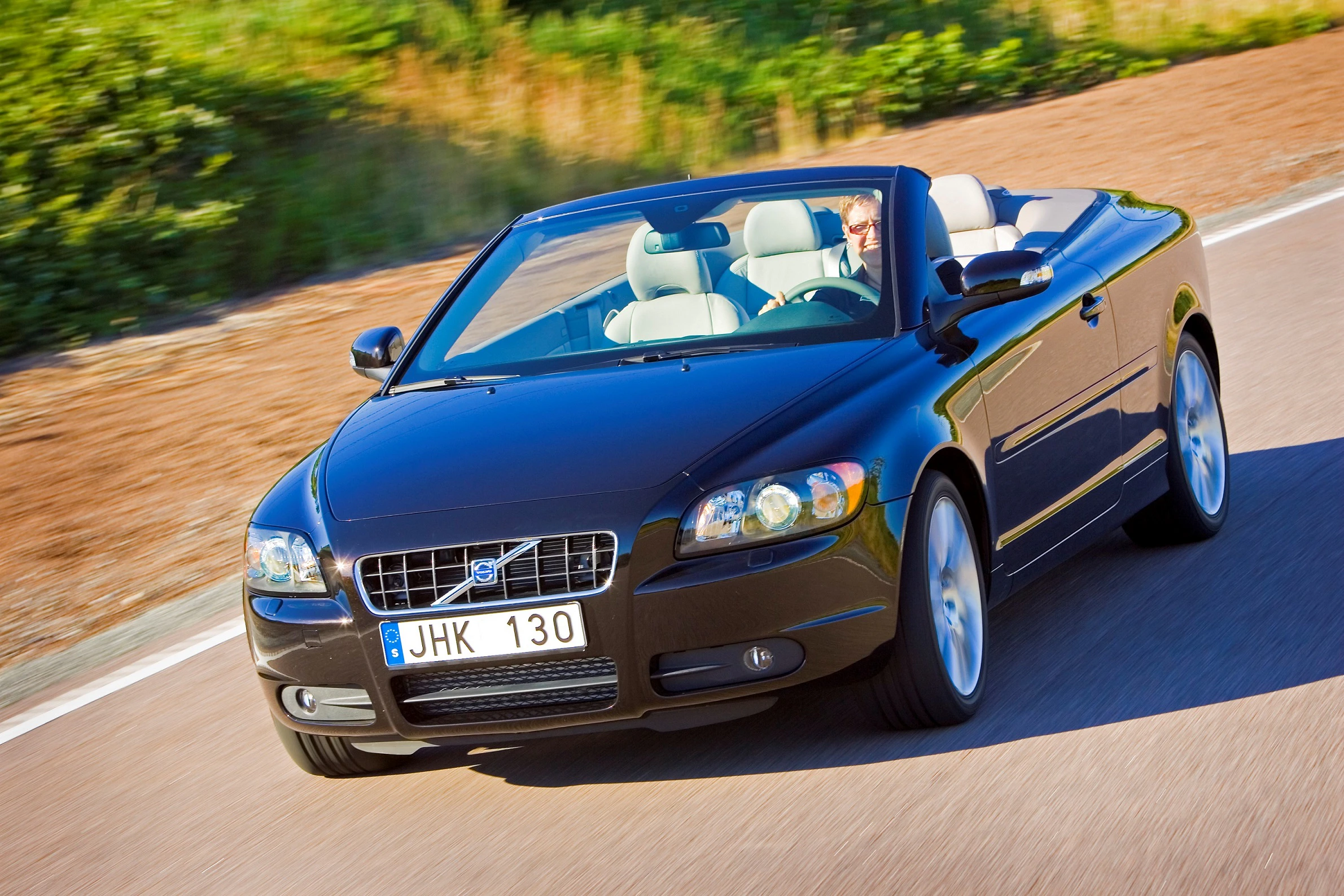 Schwarzes Cabriolet fährt auf einer Straße, bei sonnigem Wetter. Fahrer sitzt am Steuer. Hintergrund aus Bäumen und Grünflächen.