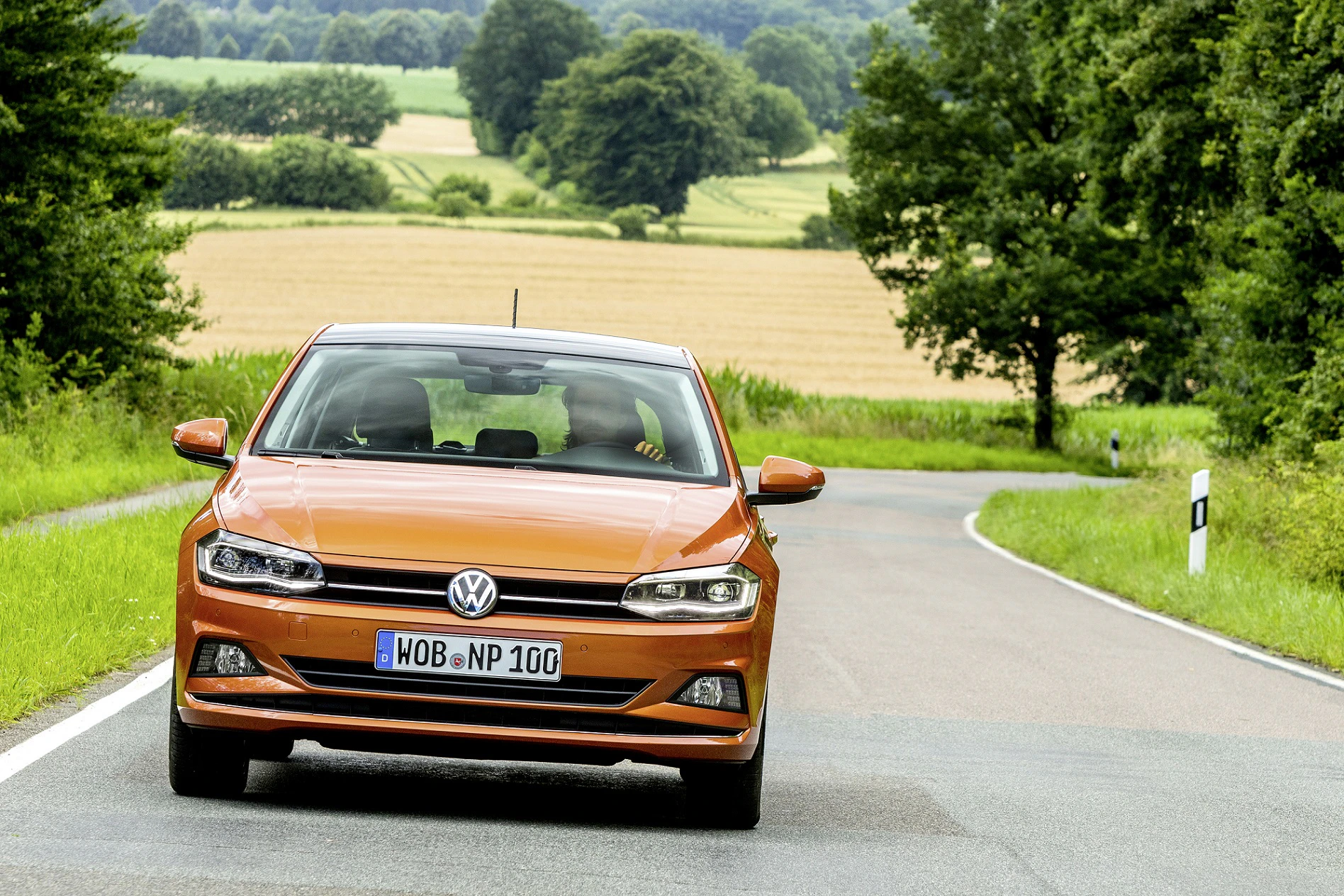 Orangefarbener Volkswagen fährt auf einer ländlichen Straße, umgeben von Bäumen und Feldern bei bewölktem Himmel.