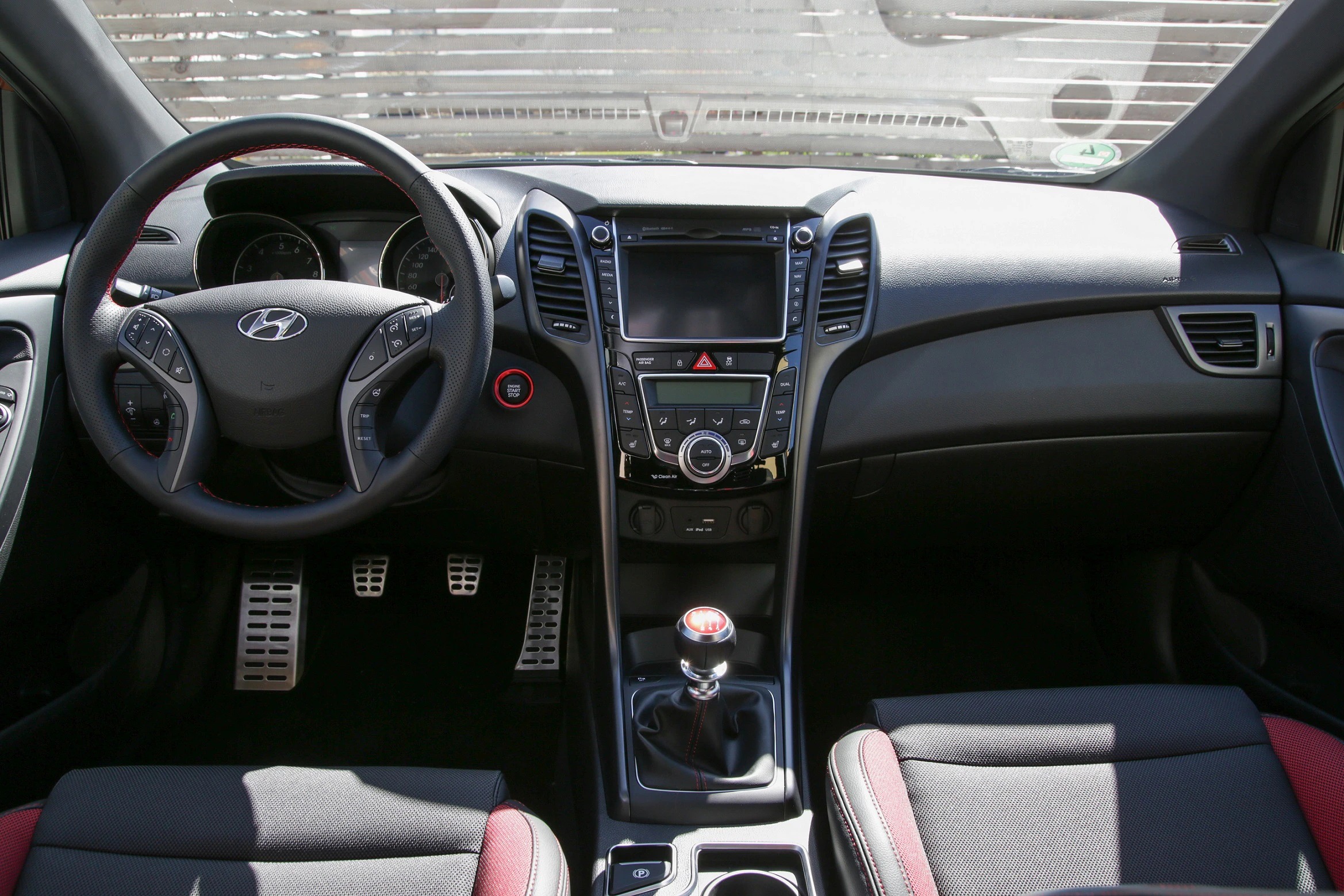 Innenraum eines Autos mit schwarzem und rotem Interieur, Lenkrad links, Schalthebel in der Mitte und Multimedia-Bildschirm in der Mittelkonsole.