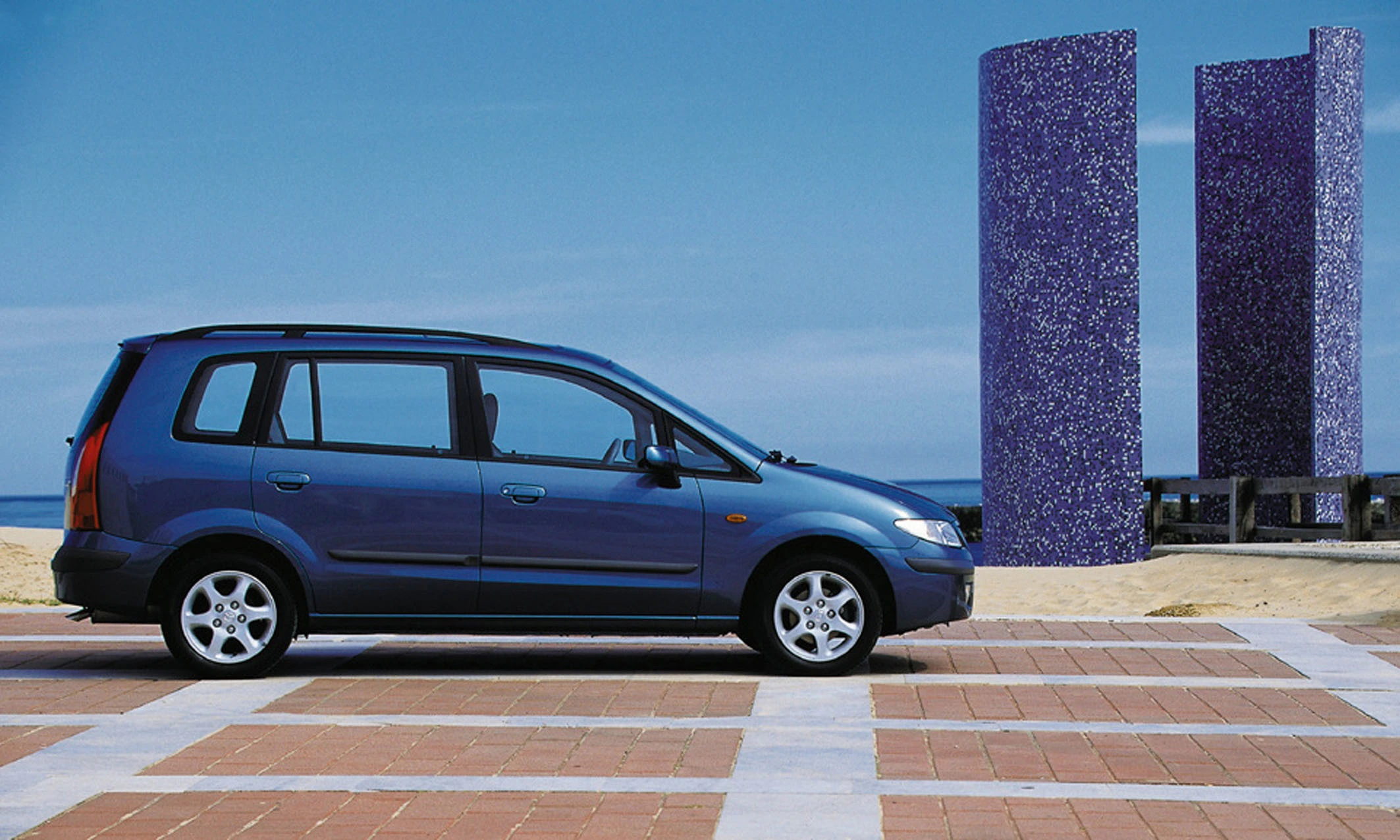 Blaues Auto parkt auf gepflastertem Platz am Strand, mit zwei violetten Skulpturen und blauem Himmel im Hintergrund.