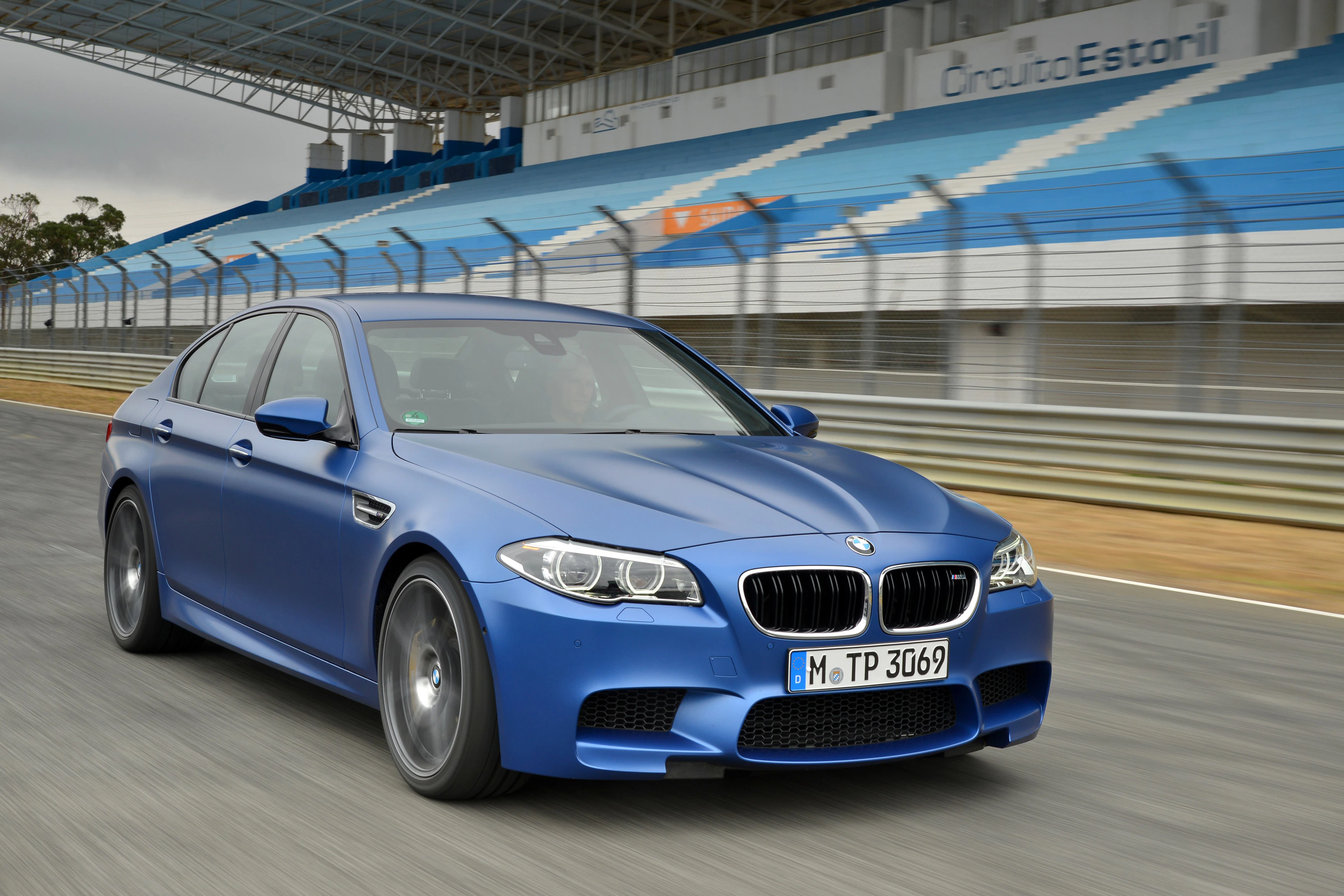 Blauer BMW fährt auf Rennstrecke mit leerer Tribüne im Hintergrund. Sportlicher Look, grauer Himmel, dynamische Bewegung.