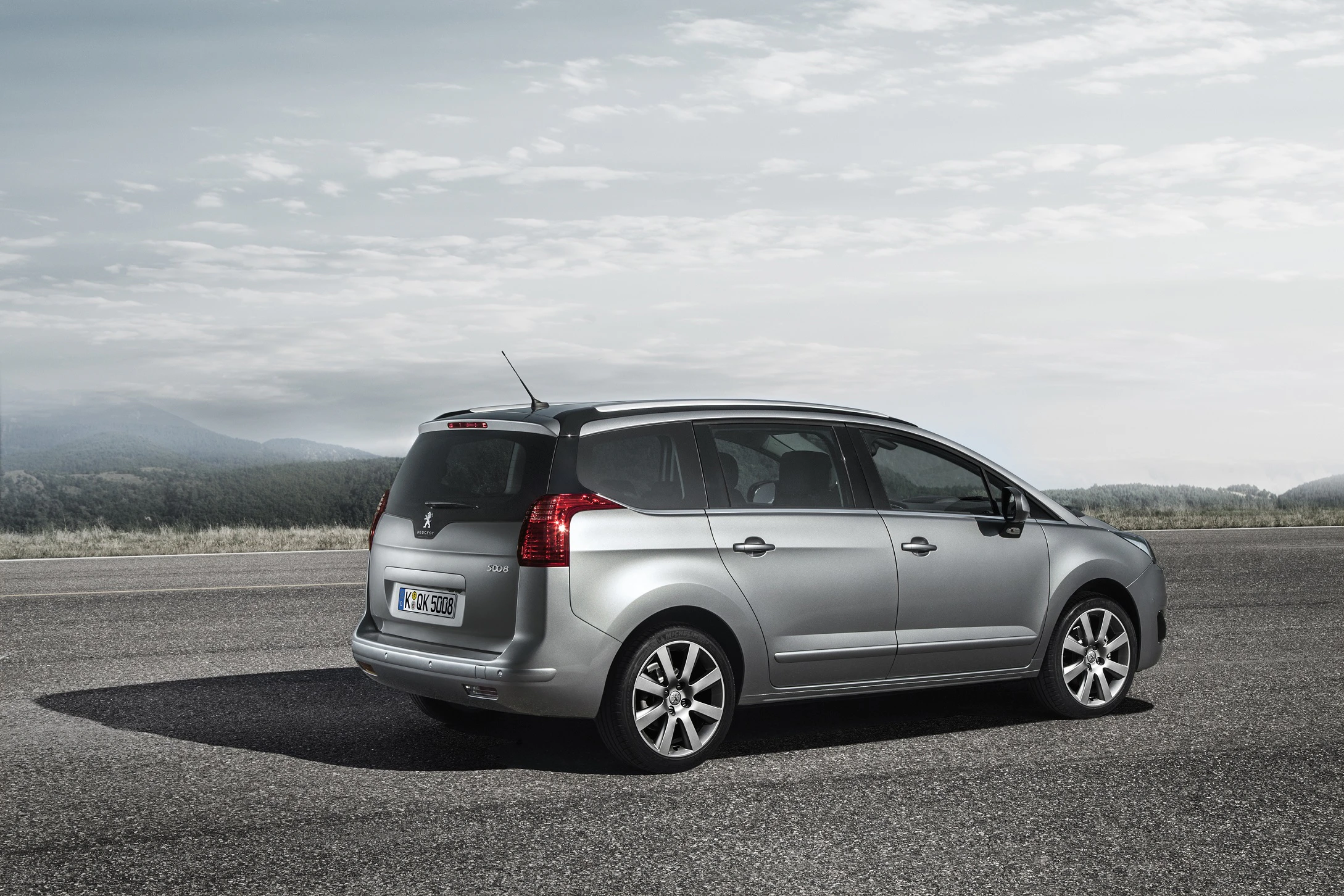 Silberner Peugeot 5008 parkt auf einer Landstraße vor bewölktem Himmel und Waldlandschaft im Hintergrund.
