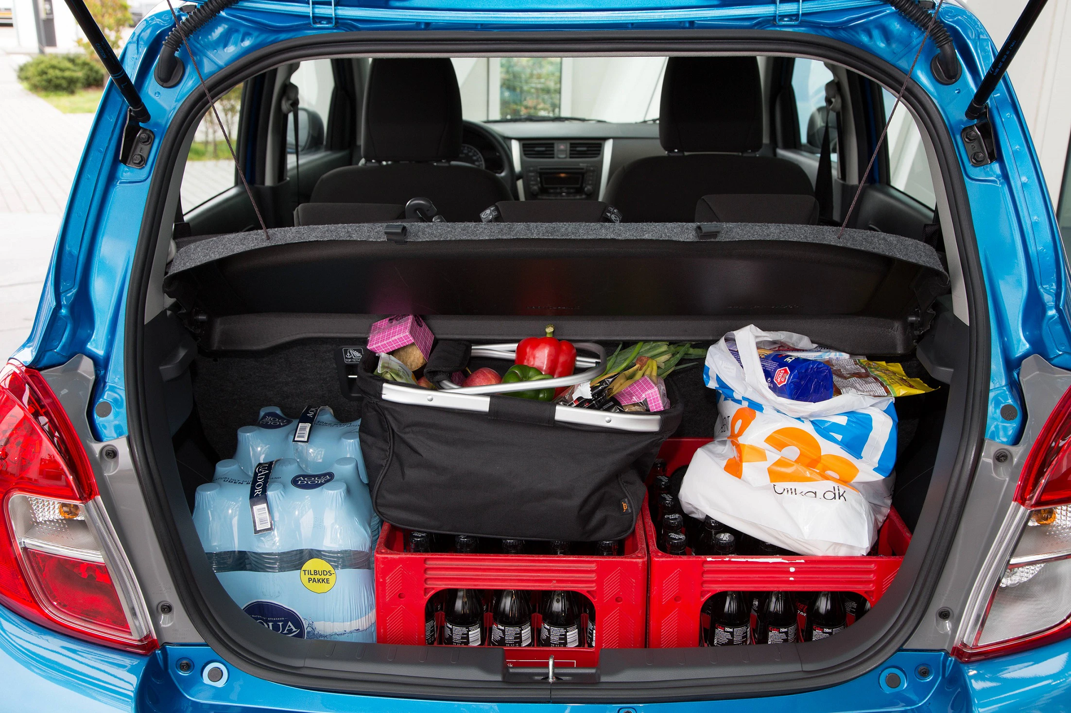 Geöffneter Kofferraum eines Autos, gefüllt mit Kisten voller Flaschen, Wasserkästen und Einkaufstüten. Verschiedene Lebensmittel sind sichtbar.