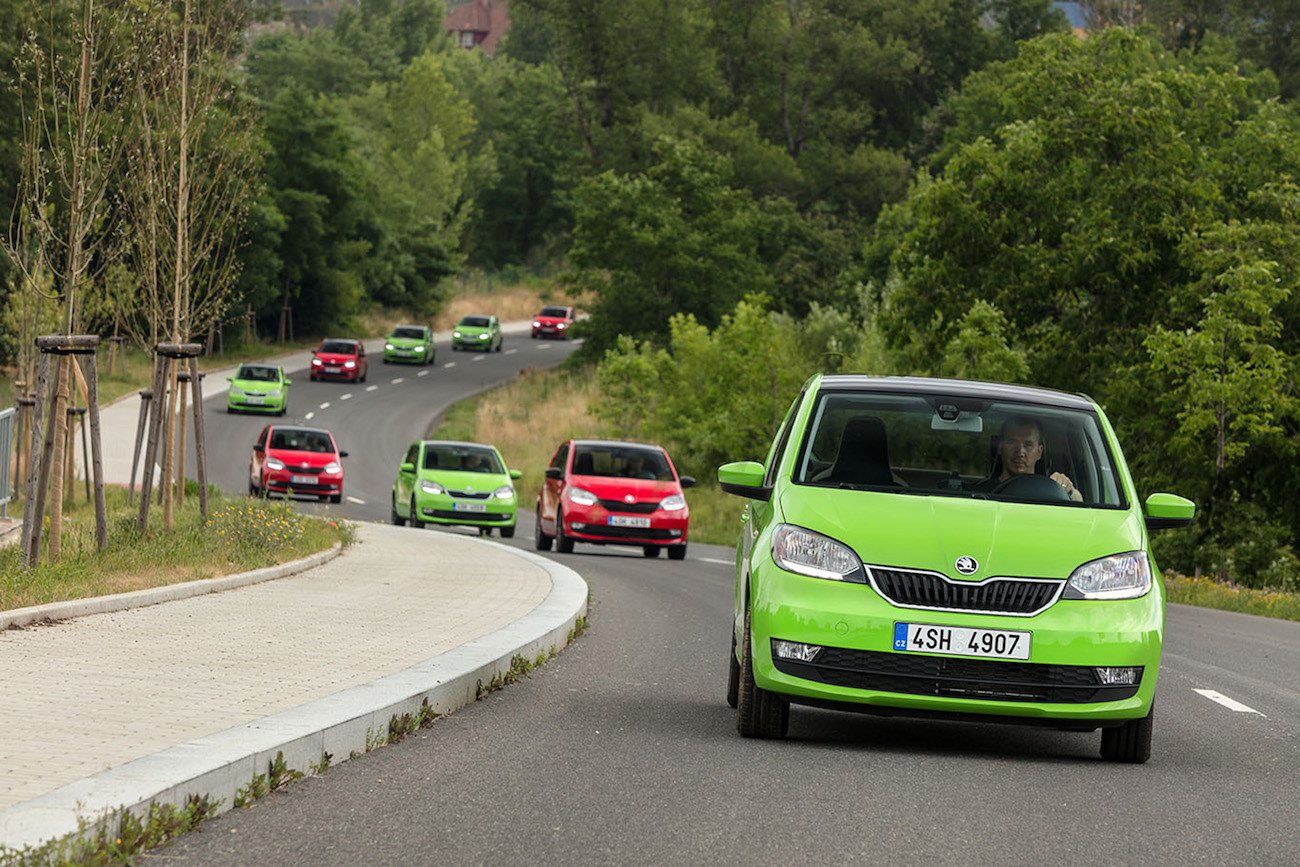 Grüne Autos fahren hintereinander auf einer kurvigen Landstraße, umgeben von Bäumen und üppiger Vegetation.