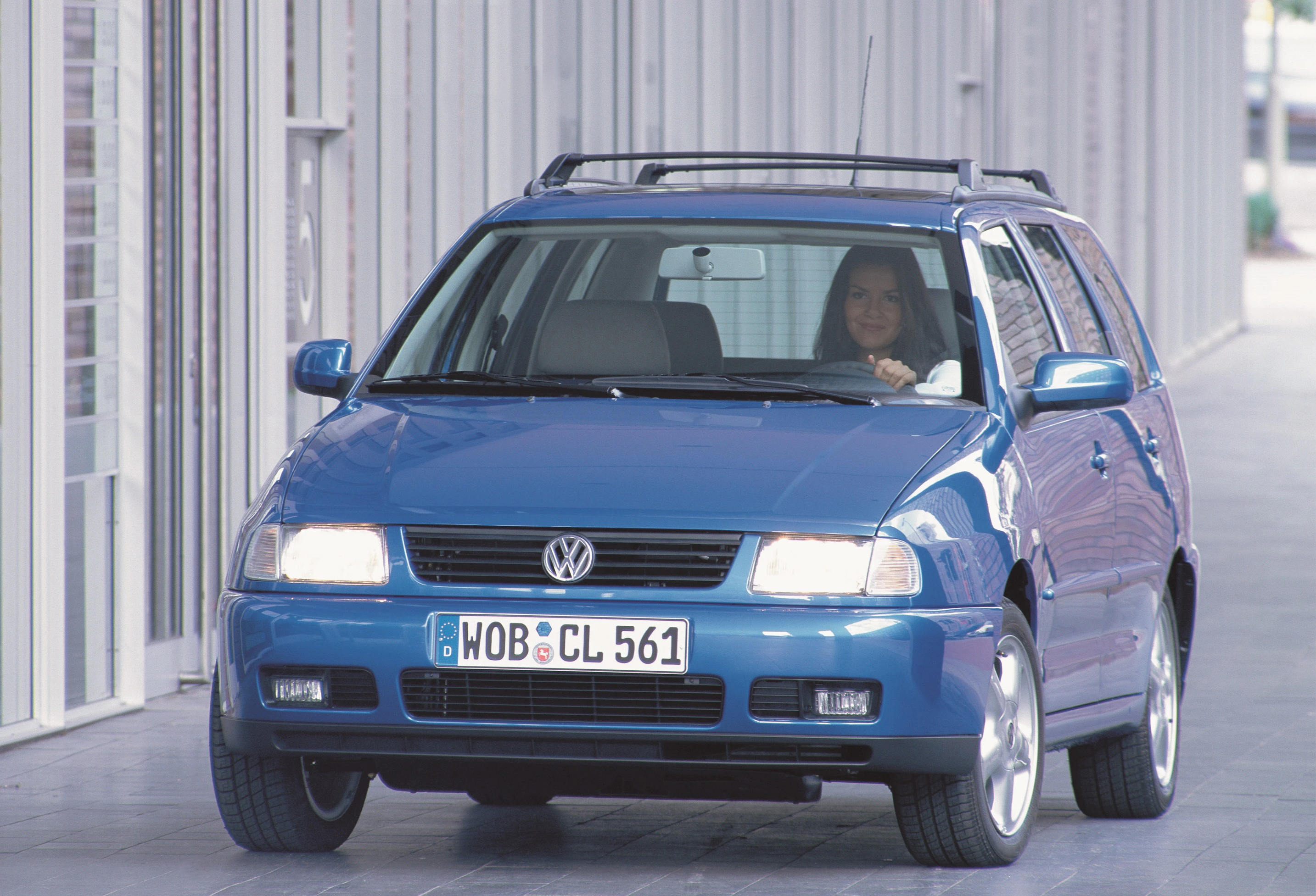 Blauer Volkswagen Kombi fährt auf einer Straße. Eine Person sitzt am Steuer. Gebäude im Hintergrund.