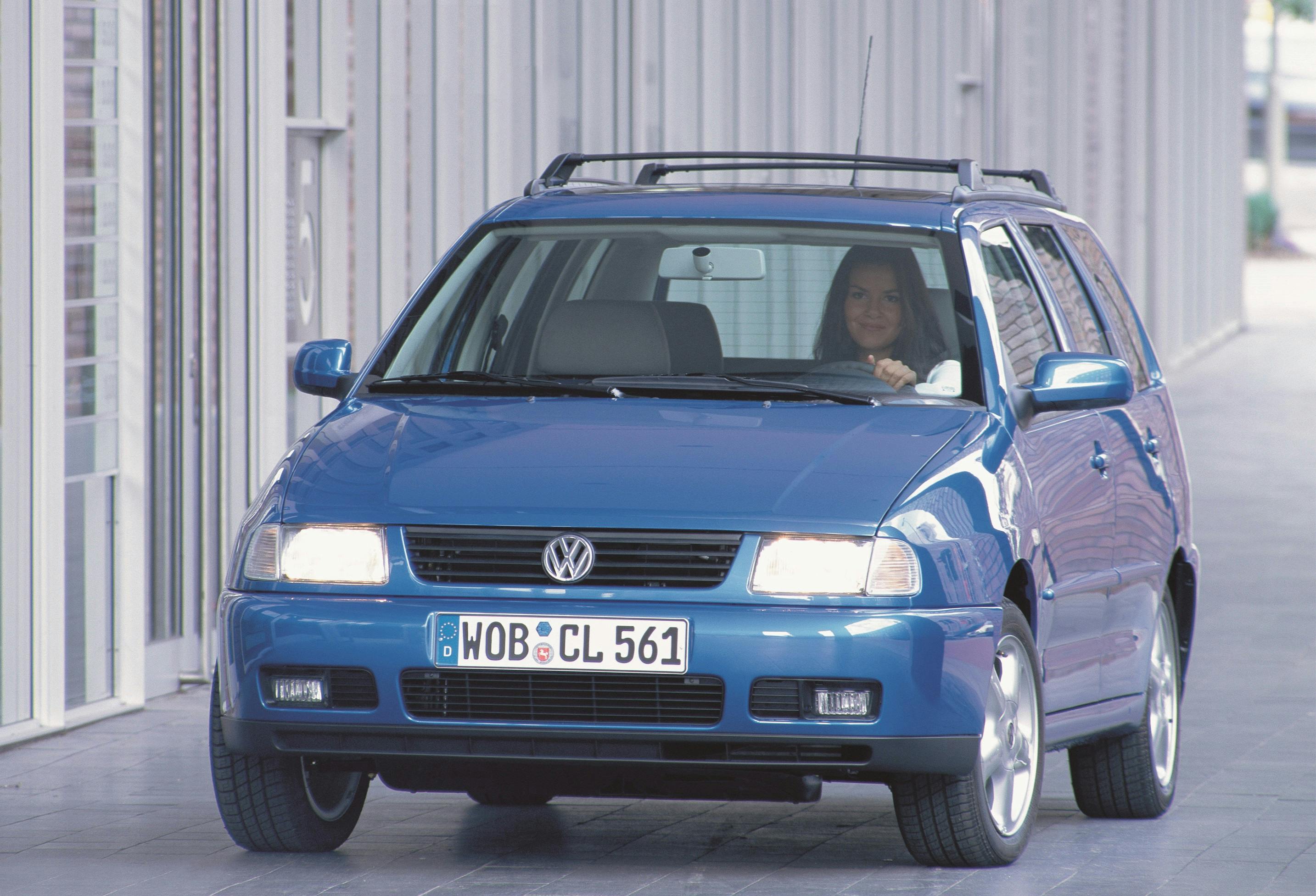 Blauer Volkswagen Kombi fährt auf einer Straße. Eine Person sitzt am Steuer. Gebäude im Hintergrund.
