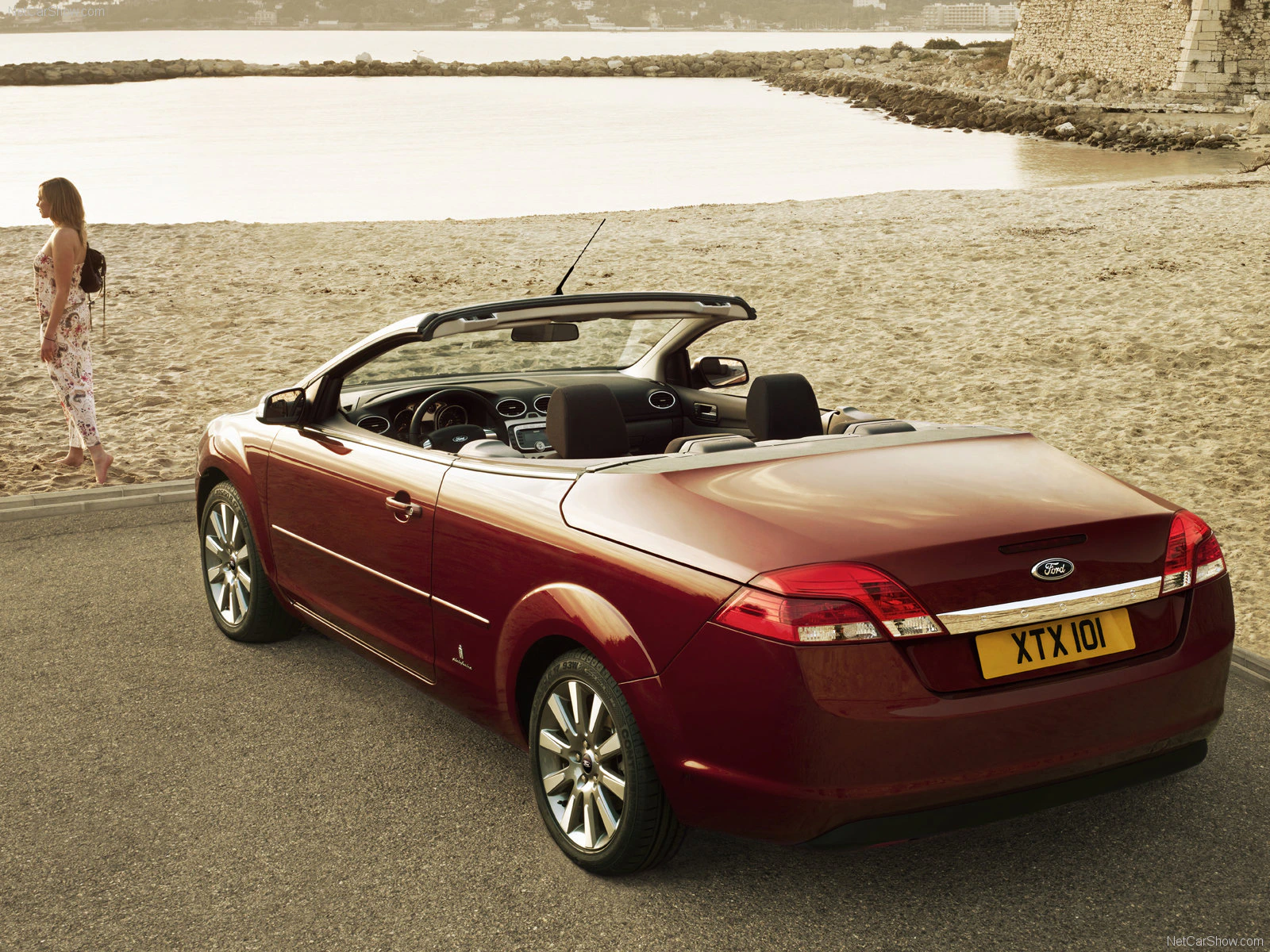 Roter Cabrio mit offenem Verdeck steht an einer Strandpromenade. Eine Frau geht am Sandstrand entlang, Wasser im Hintergrund.