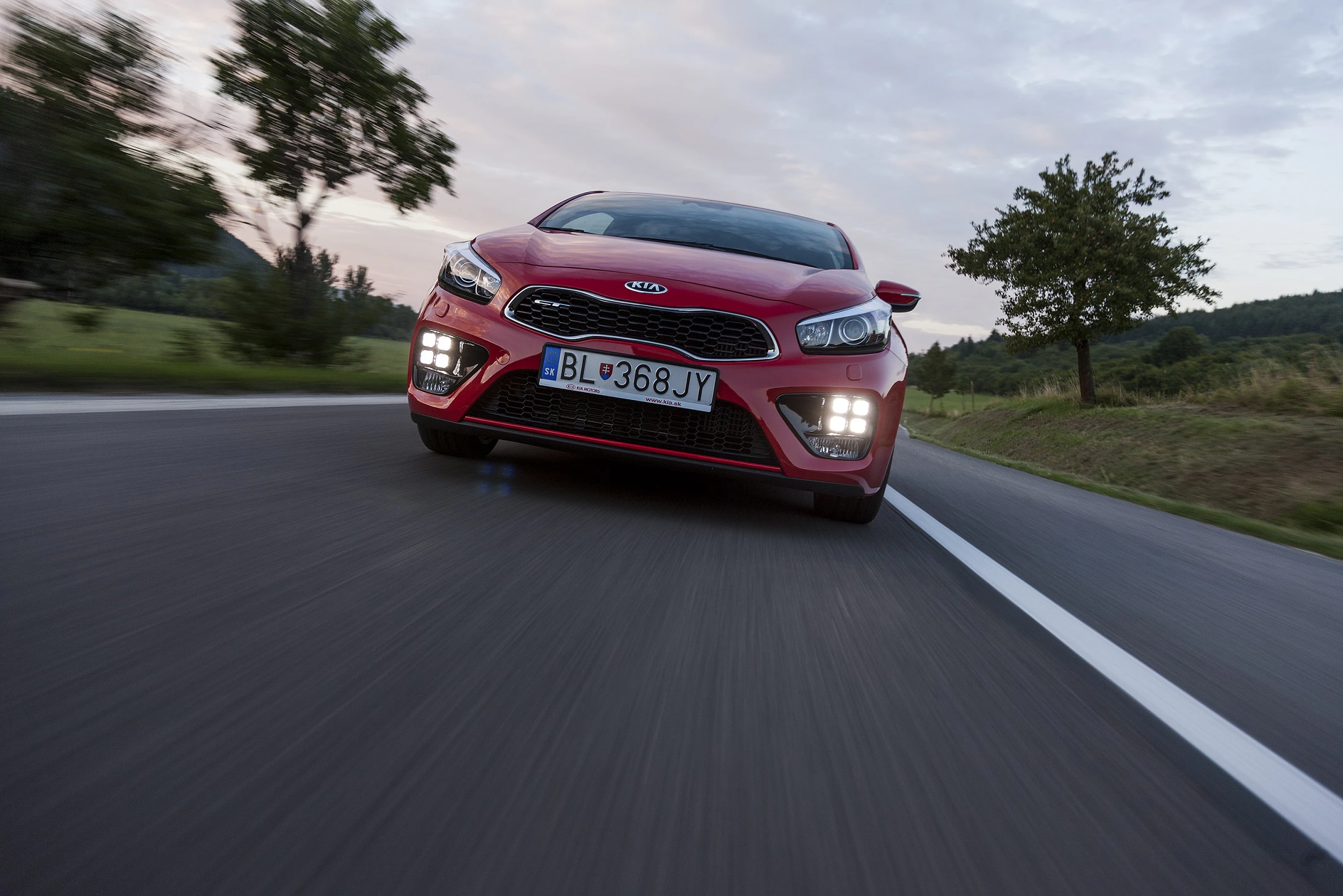 Roter Kia fährt auf Landstraße, frontal fotografiert. Hintergrund: Bäume und leicht bewölkter Himmel.