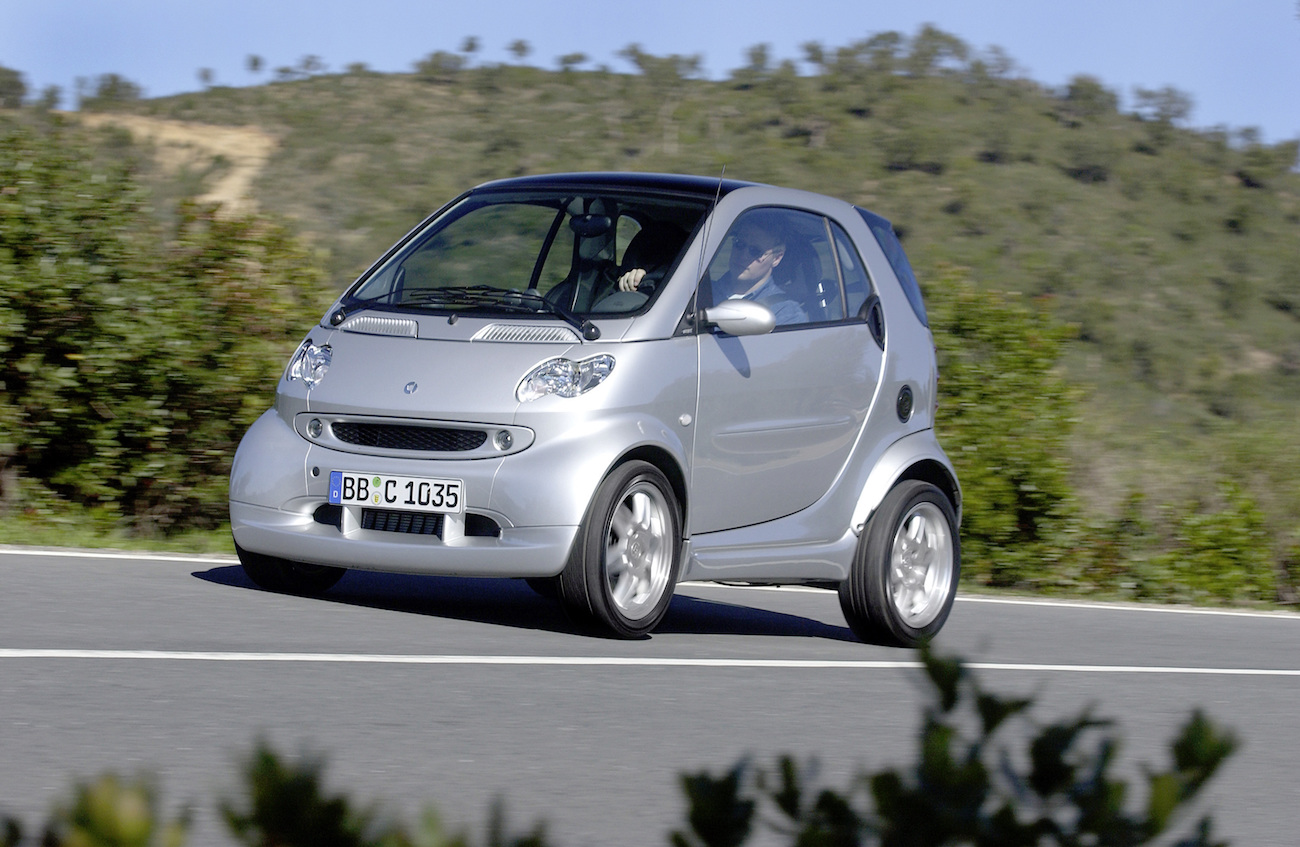 Silberner Smart Fortwo fährt auf einer Landstraße, umgeben von grüner Vegetation und Hügeln im Hintergrund.