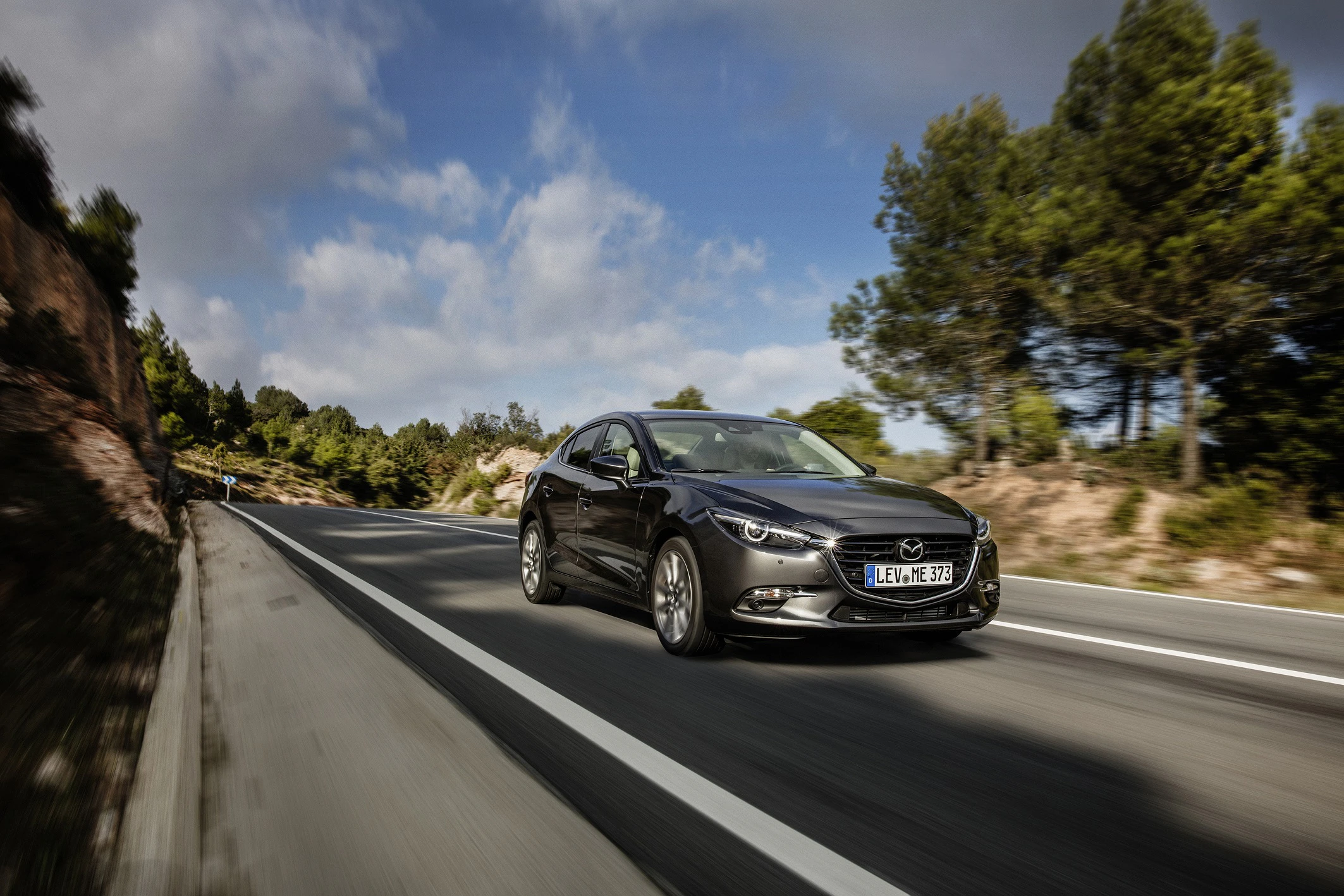 Schwarzer Mazda fährt auf einer Landstraße, umgeben von Bäumen und blauem Himmel mit Wolken.