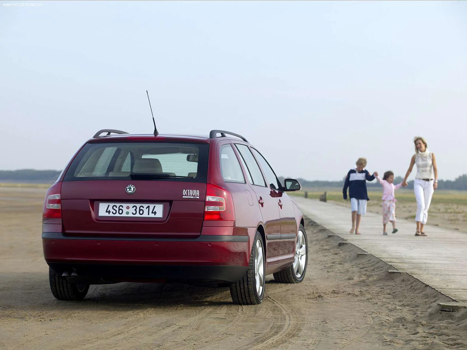 Roter Kombi parkt auf sandigem Gelände. Im Hintergrund gehen eine Frau und drei Kinder auf einem Holzsteg spazieren.